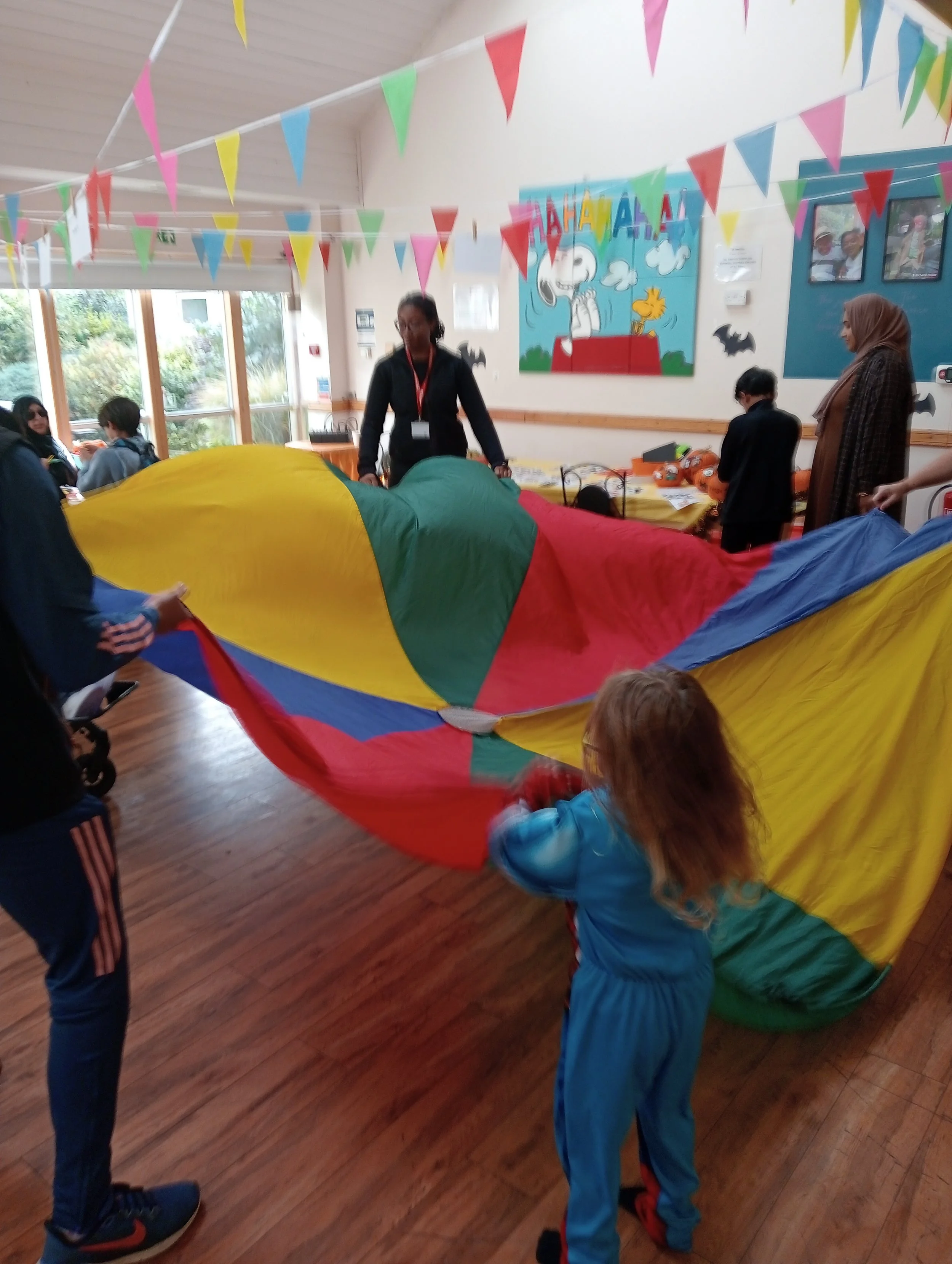 Children and adults playing with a colorful parachute indoors, with decorations hanging from the ceiling and a Snoopy comic strip on the wall.