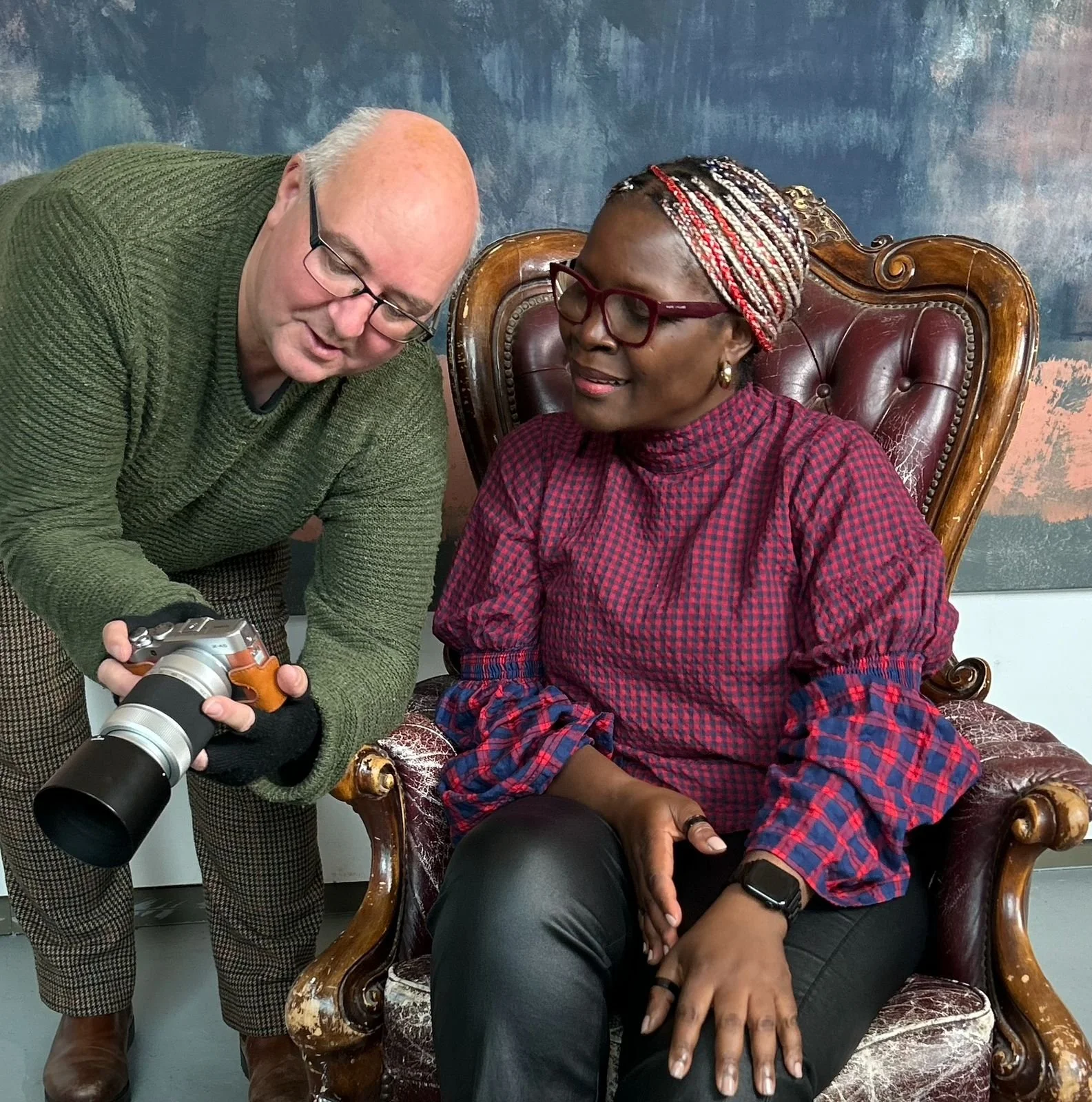 A man showing a camera to a woman sitting in an ornate leather and wood armchair, both smiling and engaged.
