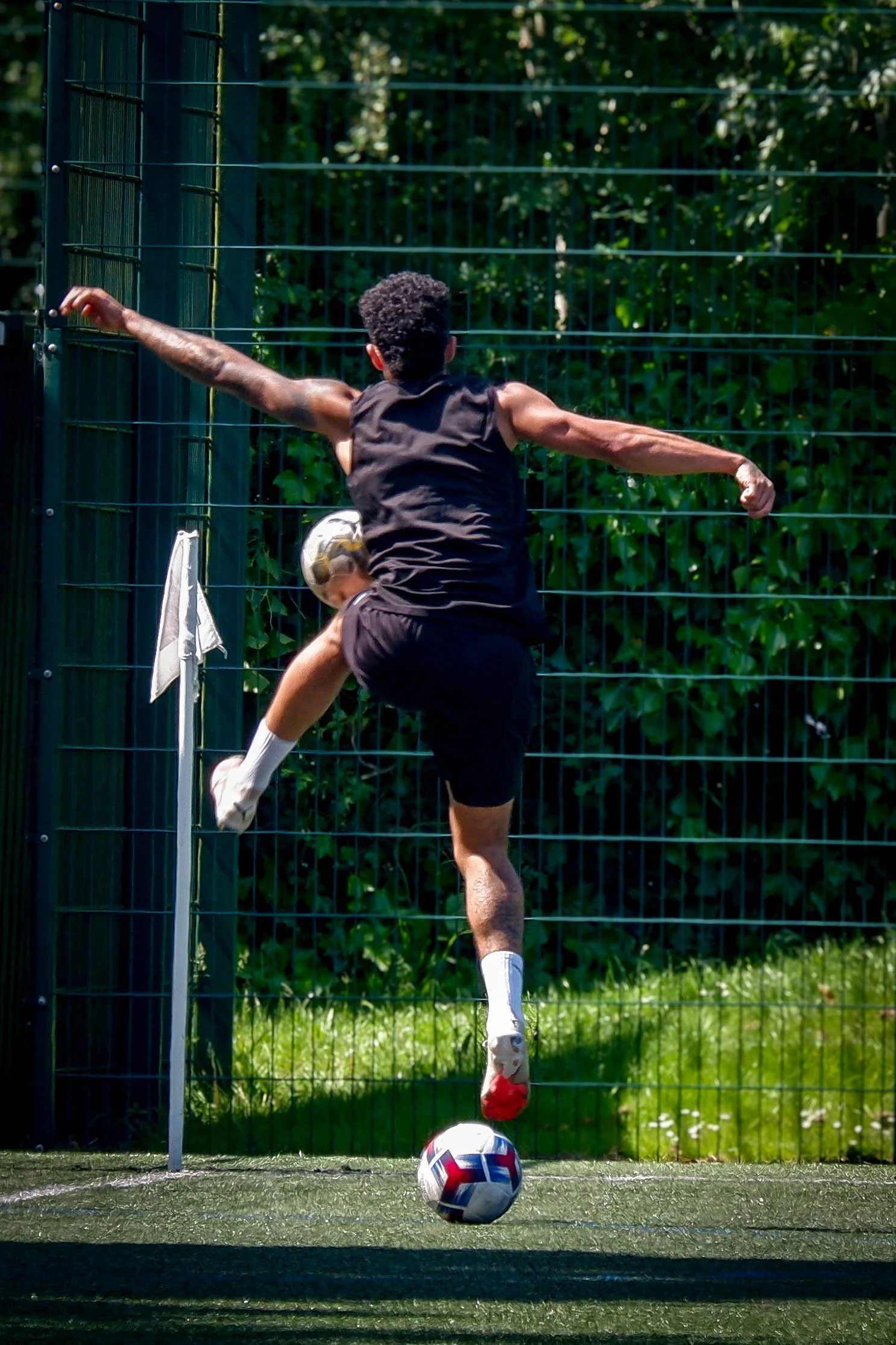 A person in black athletic clothing is jumping to kick a soccer ball on a field, with a metal fence and green trees in the background.