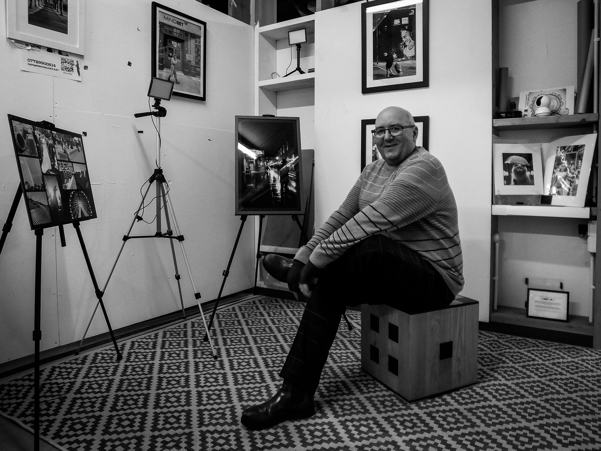 A man with glasses smiling, sitting in an art gallery surrounded by photographs and artwork on walls and on easels, with a patterned floor.