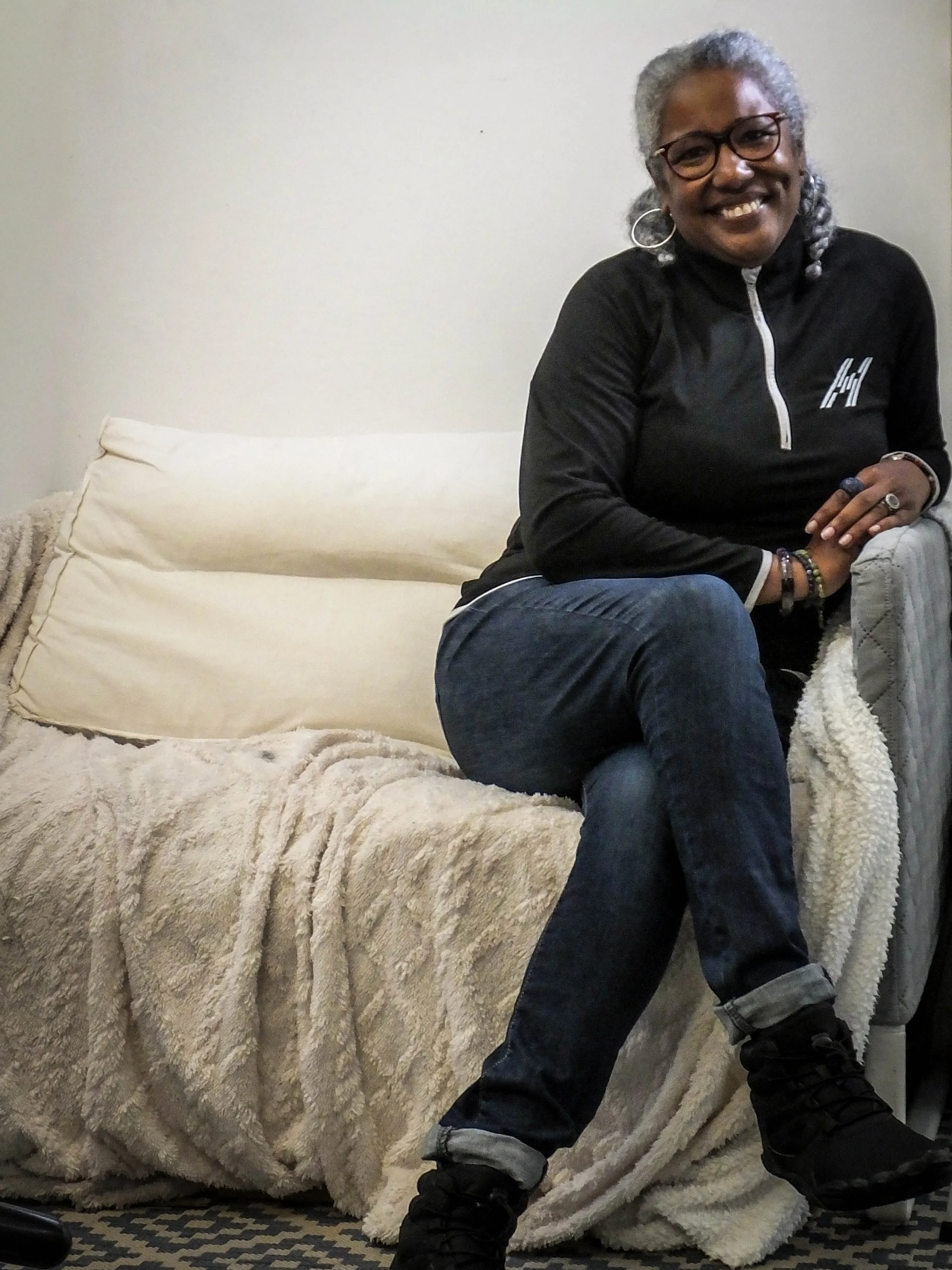 A smiling woman with gray hair, glasses, earrings, wearing a black hoodie, jeans, and black boots, sitting on a beige plush blanket in front of a white wall.