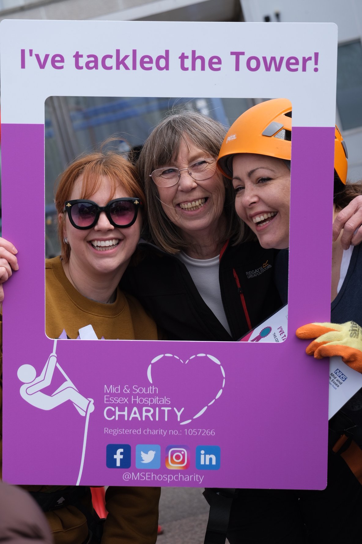 Three women smiling and holding a purple charity sign at an Essex Hospitals charity event. One woman wears sunglasses, another wears glasses, and the third wears a safety helmet.
