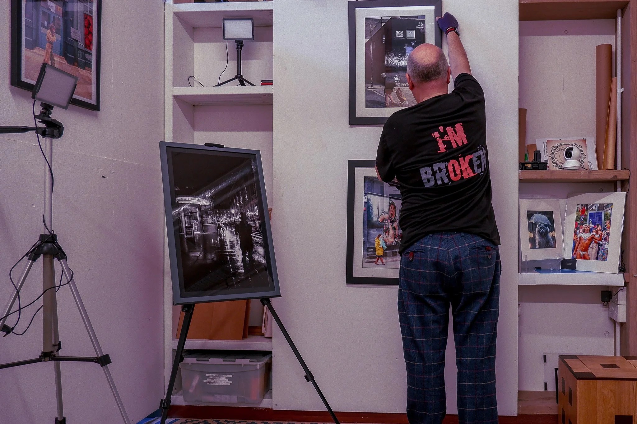 Man hanging a framed picture on a white wall in an art gallery or store, with other framed artworks and photographs displayed around.