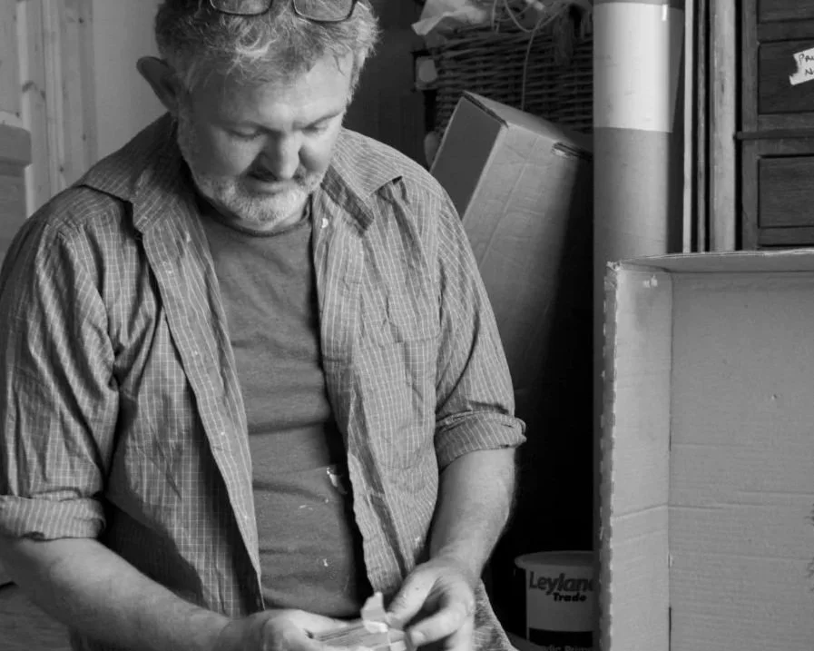 A man with gray hair and beard wearing a plaid shirt with rolled-up sleeves, looking down and holding something in his hands, in a cluttered indoor space.