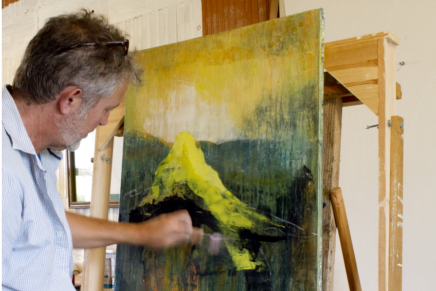 A man with glasses and a beard painting a landscape of a mountain on a canvas in a studio.
