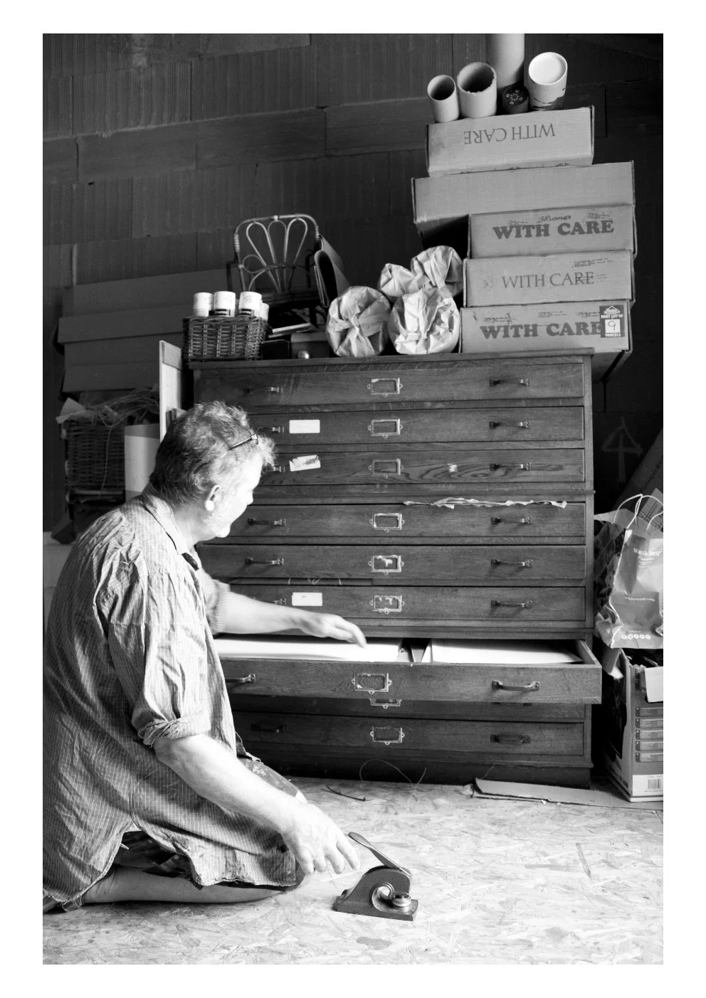 A woman kneeling on the floor, using a tape dispenser in a storage room with stacked boxes and a wooden cabinet.