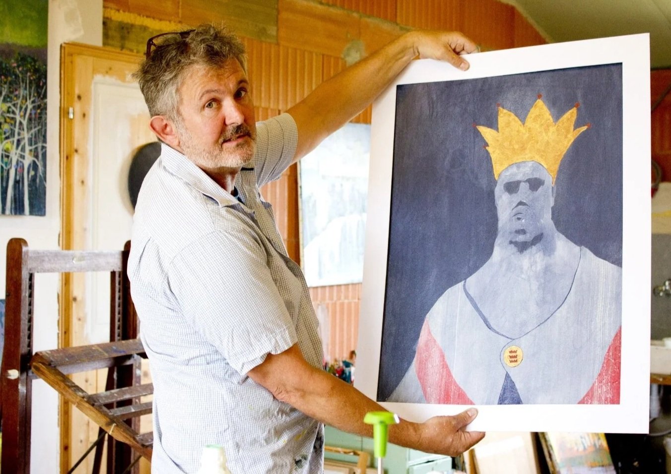 A man holding up a framed abstract painting of a person with a golden crown, wearing a shirt and a necklace, in an art studio.