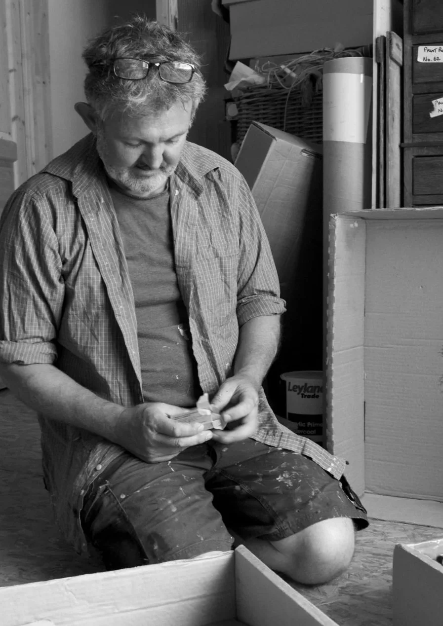 A man with glasses on his head, a beard, and casual clothing is kneeling on the floor, working with a small object in his hands. The surroundings include cardboard boxes, shelves, and cluttered storage items.