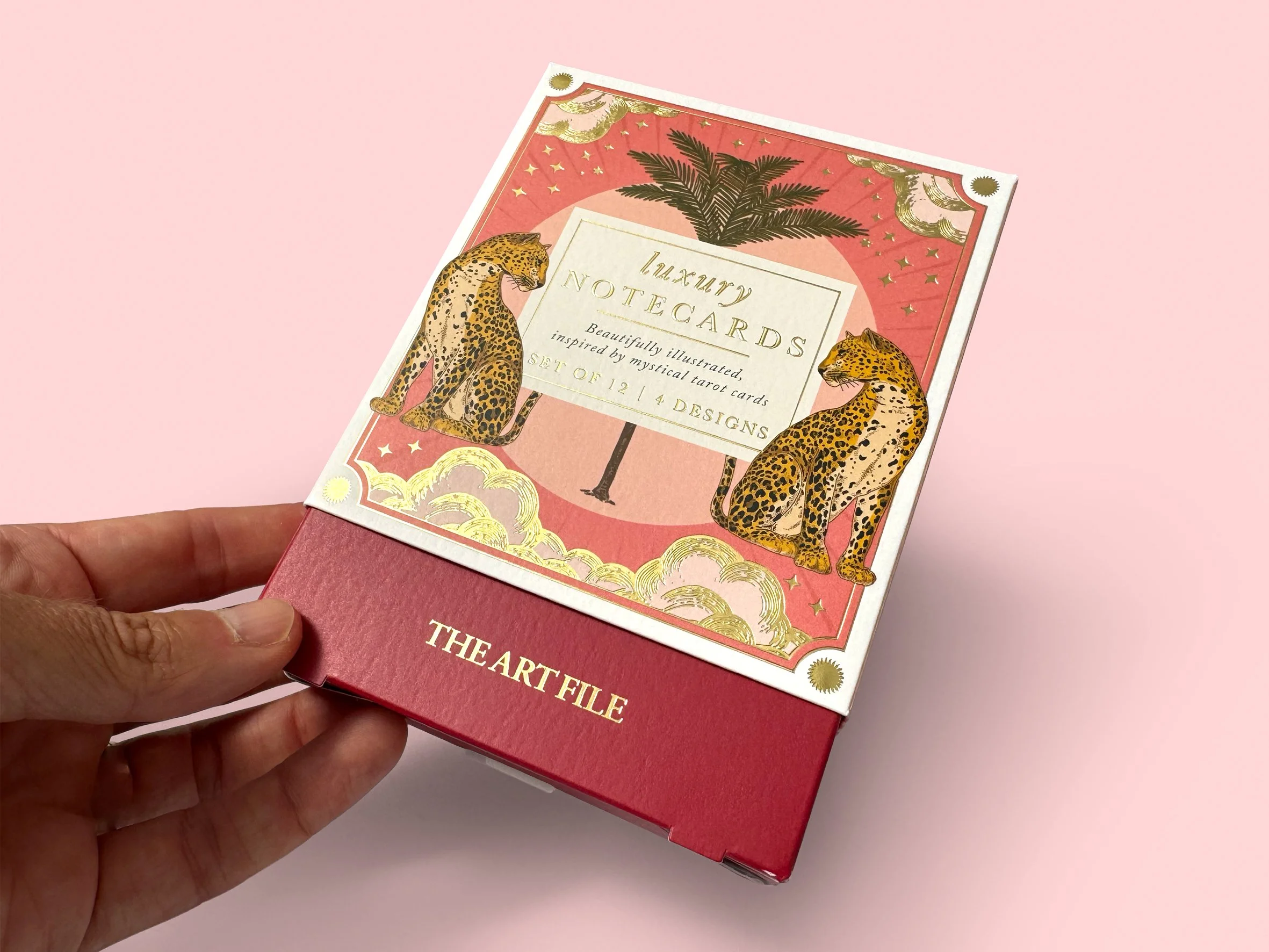 A person holding a box titled "January Notecards" with illustrations of two leopards, palm trees, gold clouds, stars, and a moon on a pink background. Carton packaging produced by Windles Packaging.