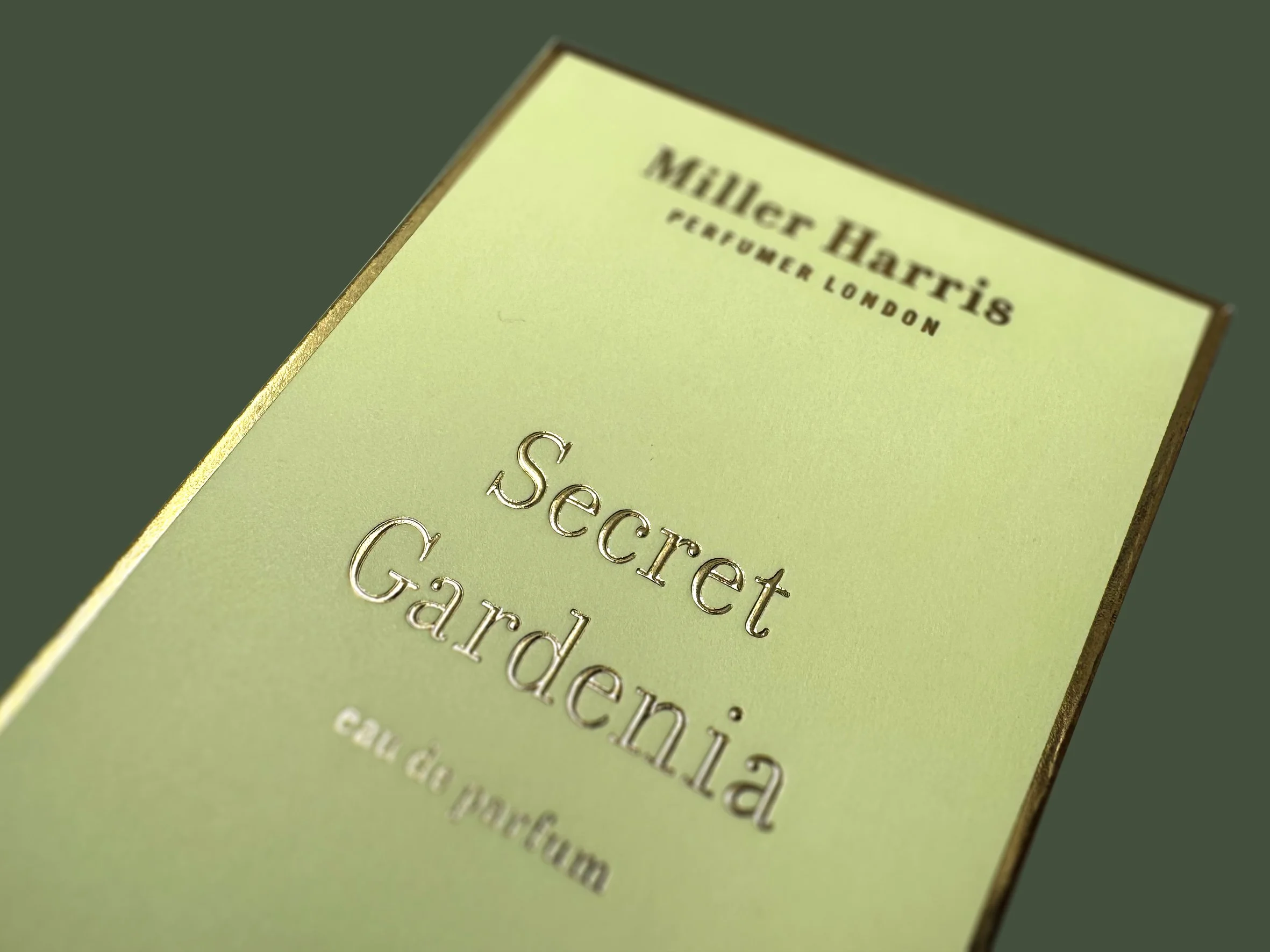 Close-up of a yellow perfume box with silver embossed text and a gold edge, reading "Miller Hartzis Perfumer London" and "Scented paste in a cardboard box."