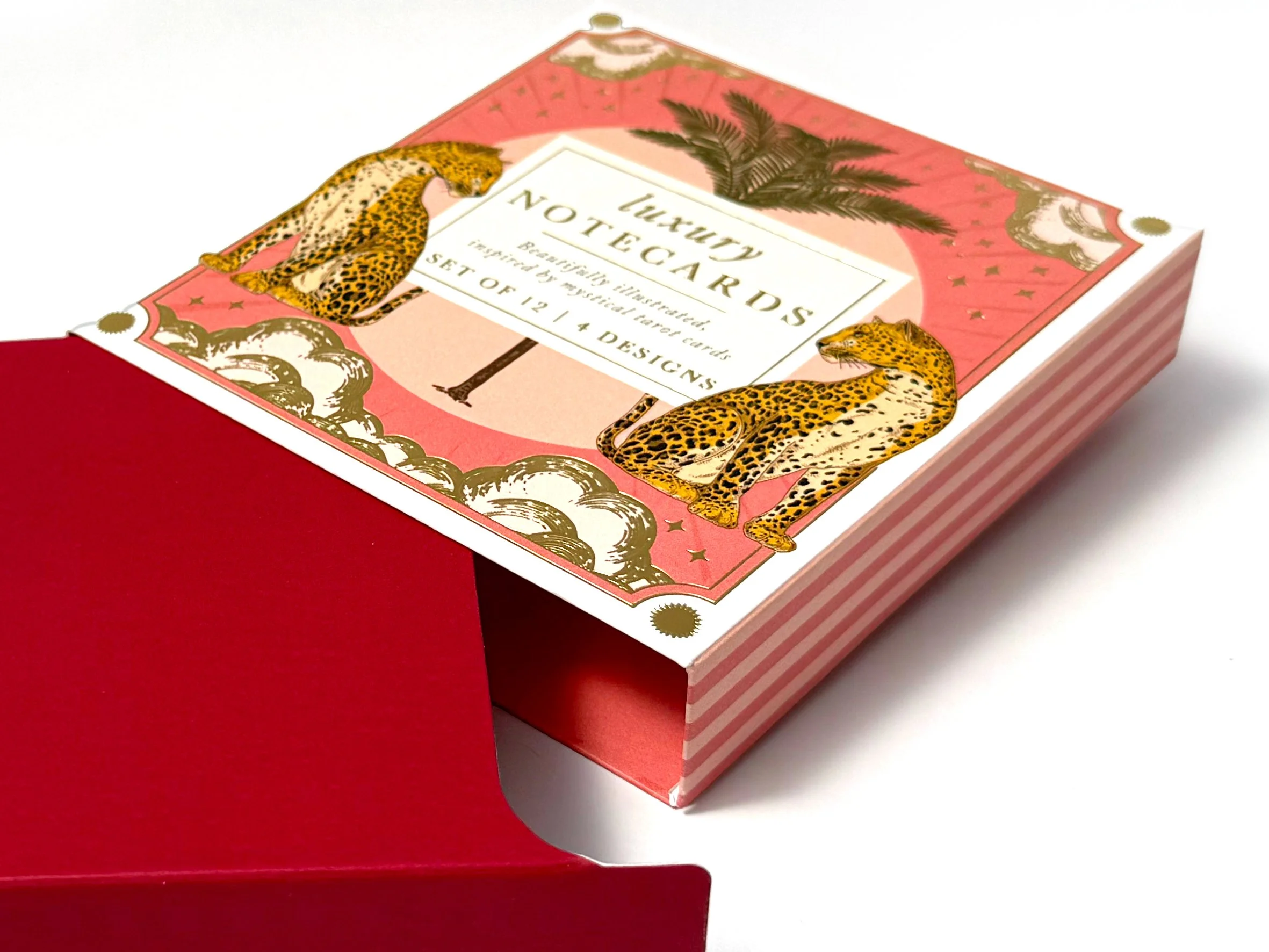 A decorative set of luxury notecards featuring illustrations of leopards, palm trees, and gold accents, partially pulled out from a pink and white striped box placed on a red surface. Carton packaging produced by Windles Packaging.