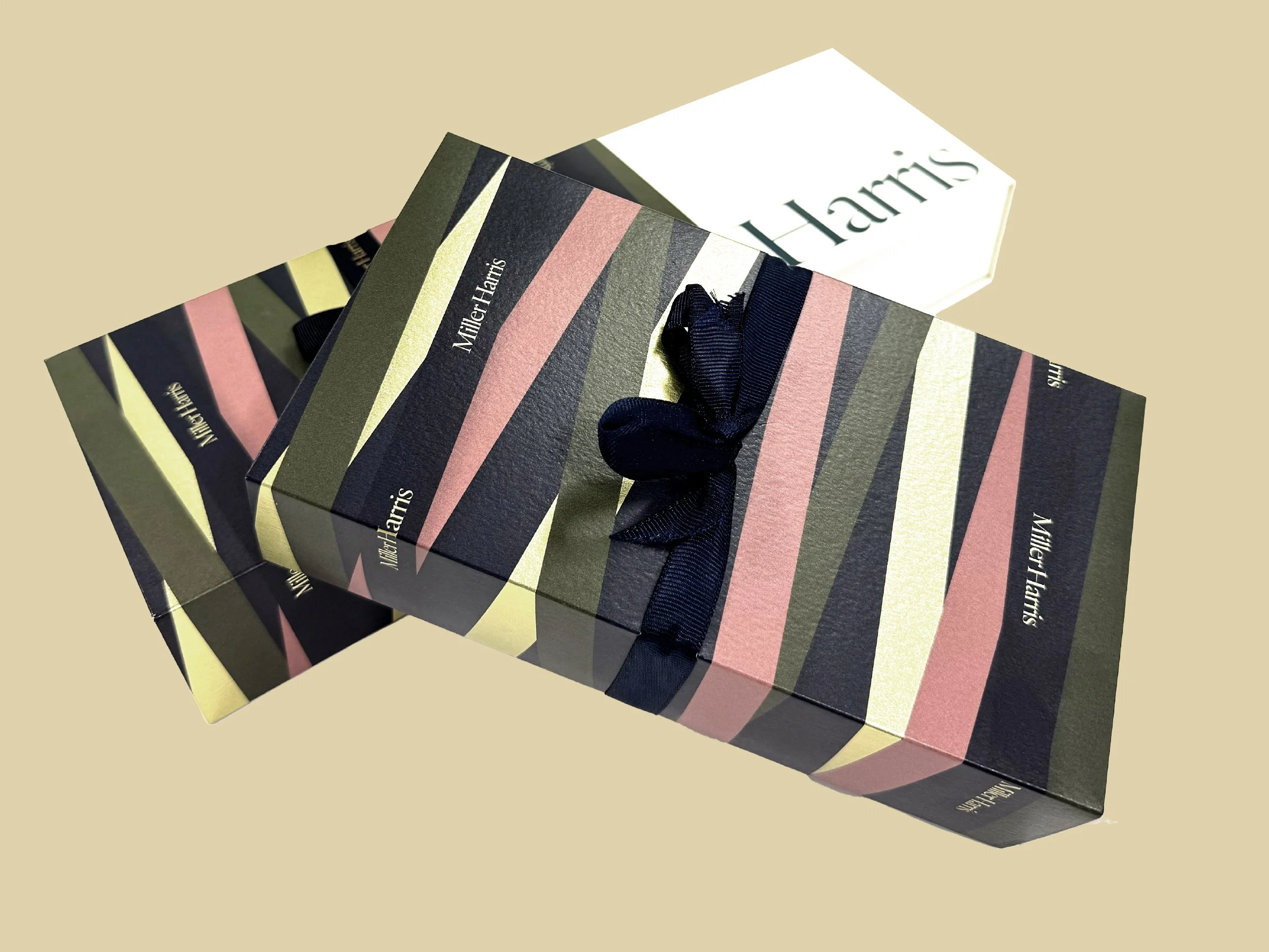 Close-up of a striped gift box with a black ribbon, featuring the name Milton Harris, and a white box with a blurred name in the background, placed on a beige surface. Carton packaging produced by Windles Packaging.