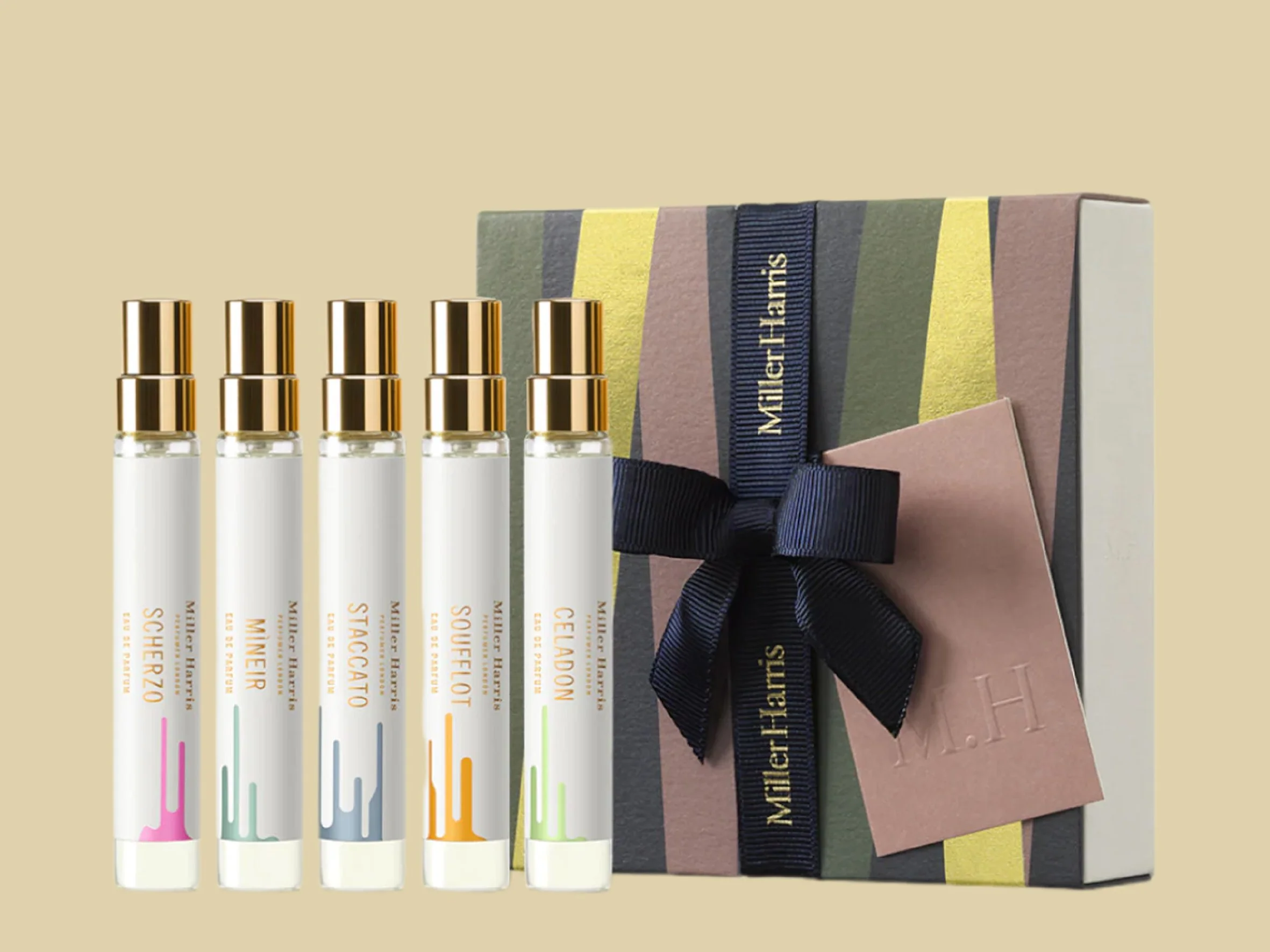 Set of six white tubes with gold caps and pastel-colored branding, arranged in front of a pastel-colored book and gift box wrapped with a black ribbon. Carton packaging produced by Windles Packaging.