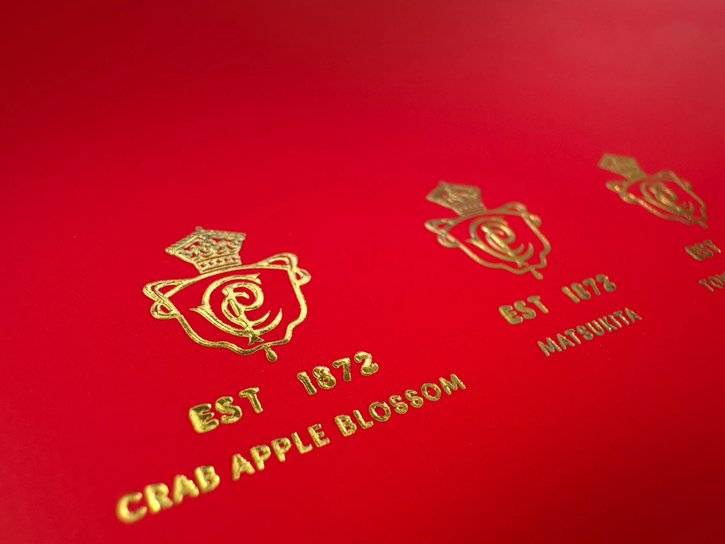 Clive Christian packaging. Close-up of a red surface with gold foil logos and text that reads "EST 1872" and "APPLE BLOSSOM". Inline cold foiling, Carton packaging produced by Windles Packaging.