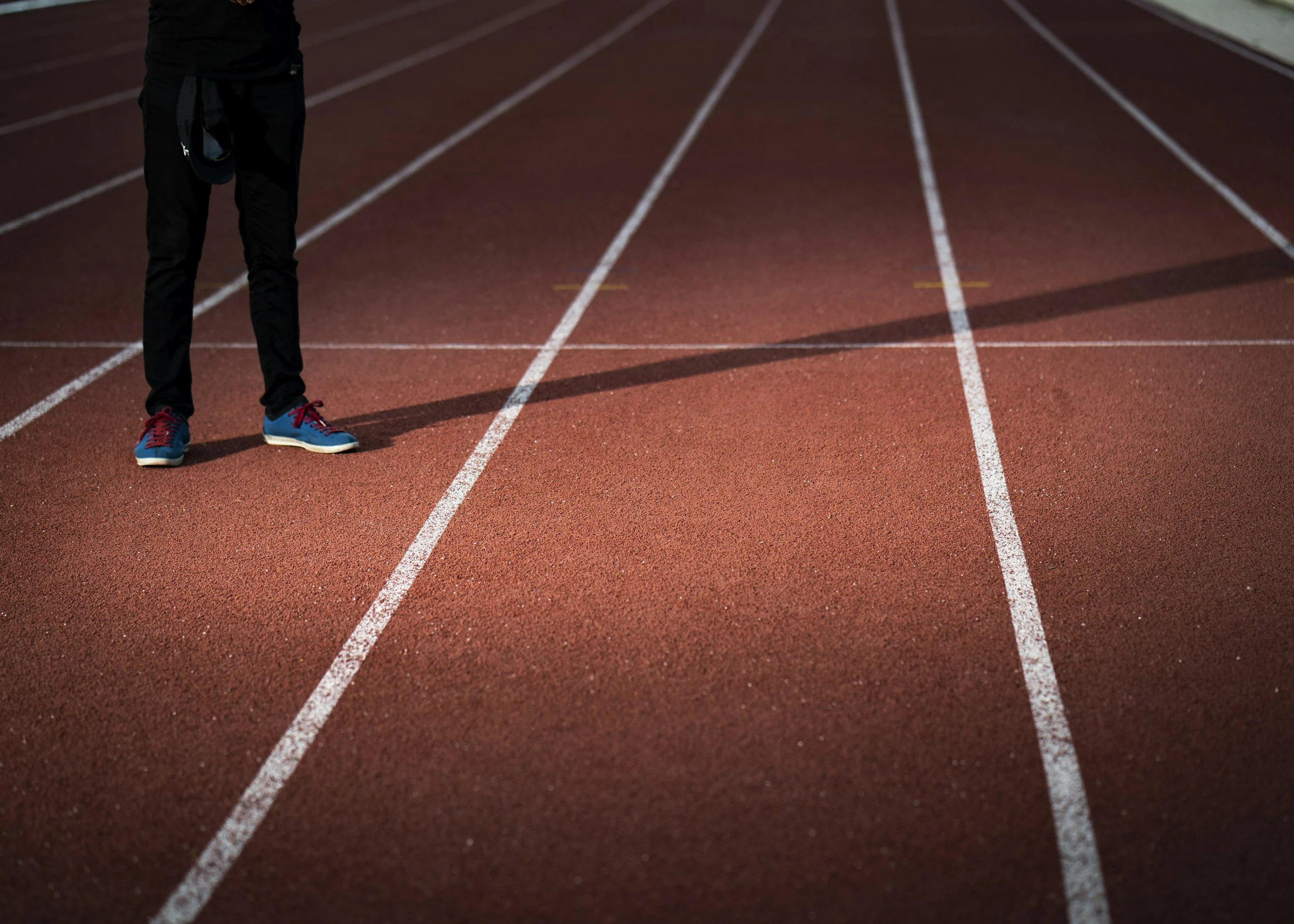A person standing on a red running track wearing black pants and blue running shoes with red laces.