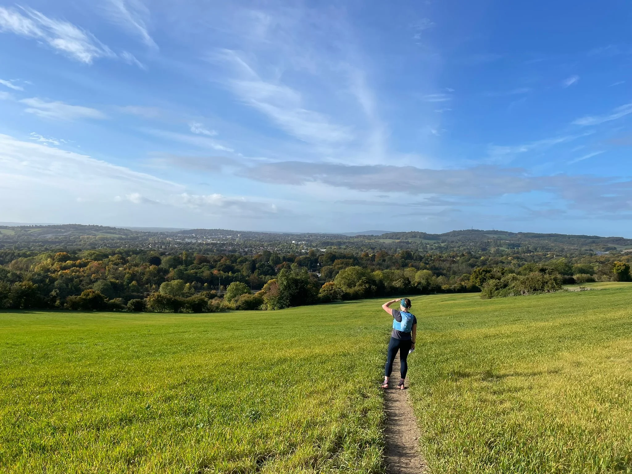 Mairi standing on a grassy trail, looking out over hills and trees under a blue sky.