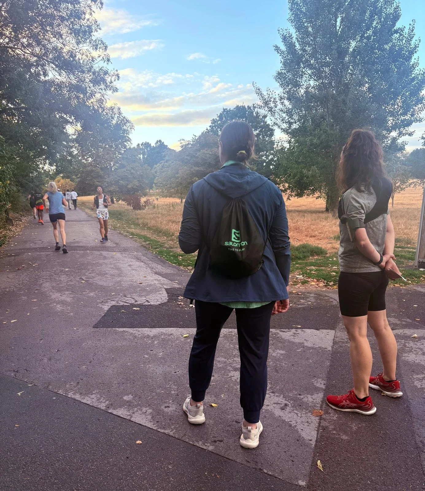 Mairi coaching runners on a tarmac path at Primrose Hill, dry grass to one side and trees beyond.