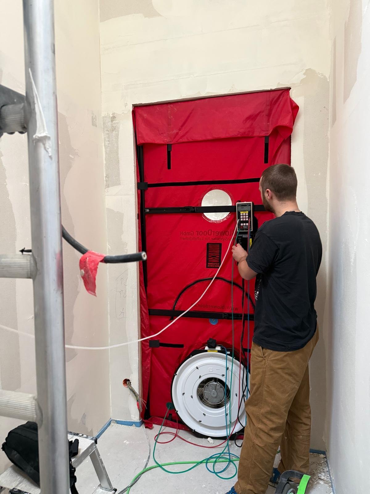 Ein Mann arbeitet an einer Installation oder Wartung eines Lüftungssystems in einem Raum unter Bauarbeiten. Er benutzt ein Messgerät, und die Wand ist teilweise unvollständig gestrichen.