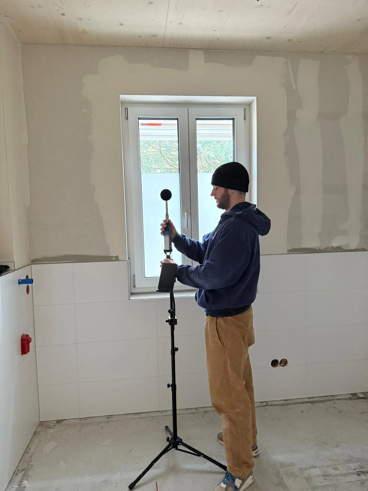 Ein Mann in einer Baustelle prüft die Bauakustik mit einem Messgerät.