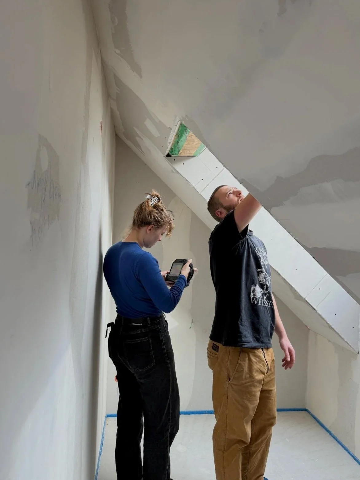 Zwei Personen, ein Mann und eine Frau, in einem Raum mit unvollständiger Decke, der Mann arbeitet an der Decke und die Frau schaut auf ihr Smartphone.