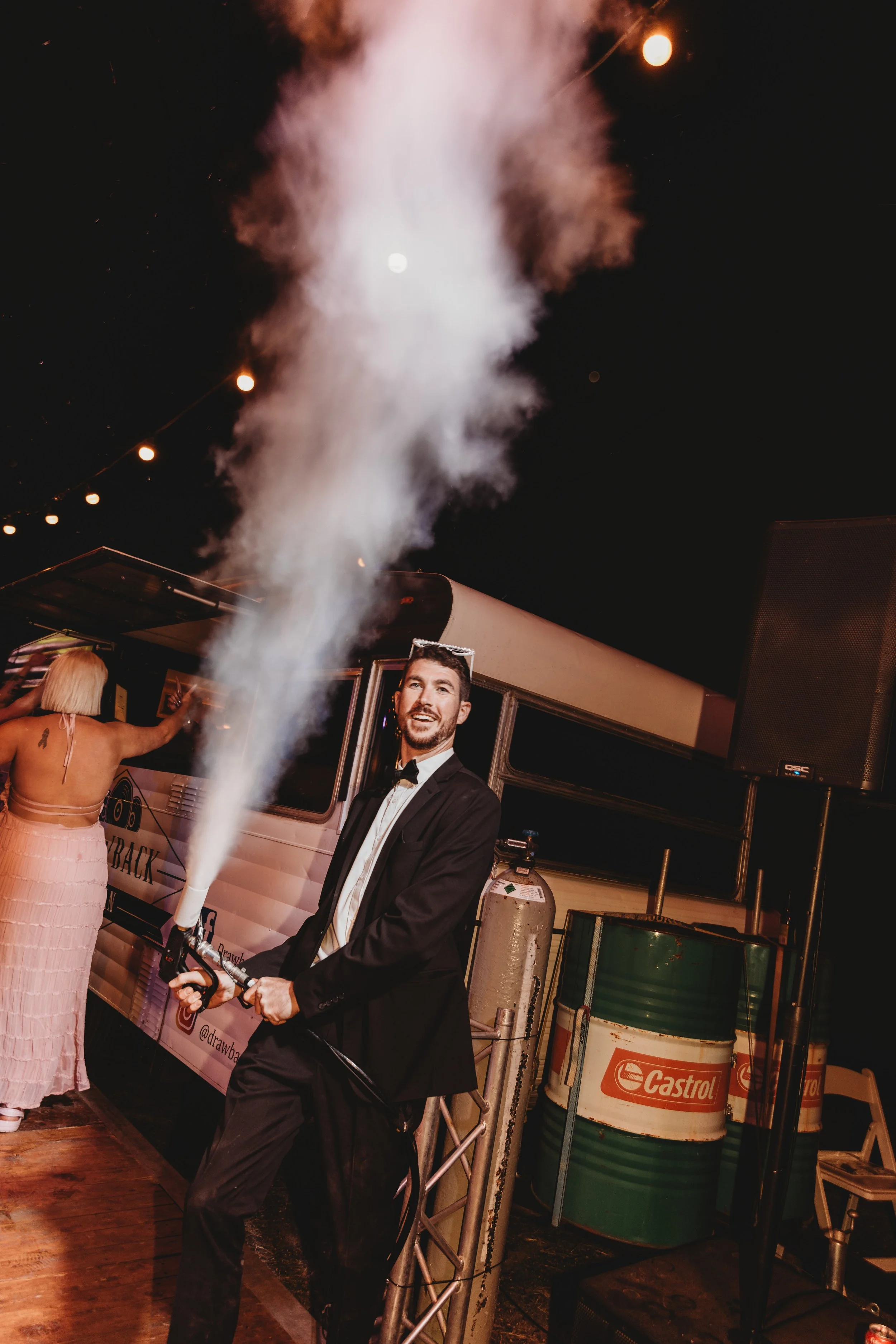 A man in a tuxedo holding a sparkler at night with fireworks and string lights in the background. A woman in a pink dress stands nearby. The setting appears to be a celebration or party.