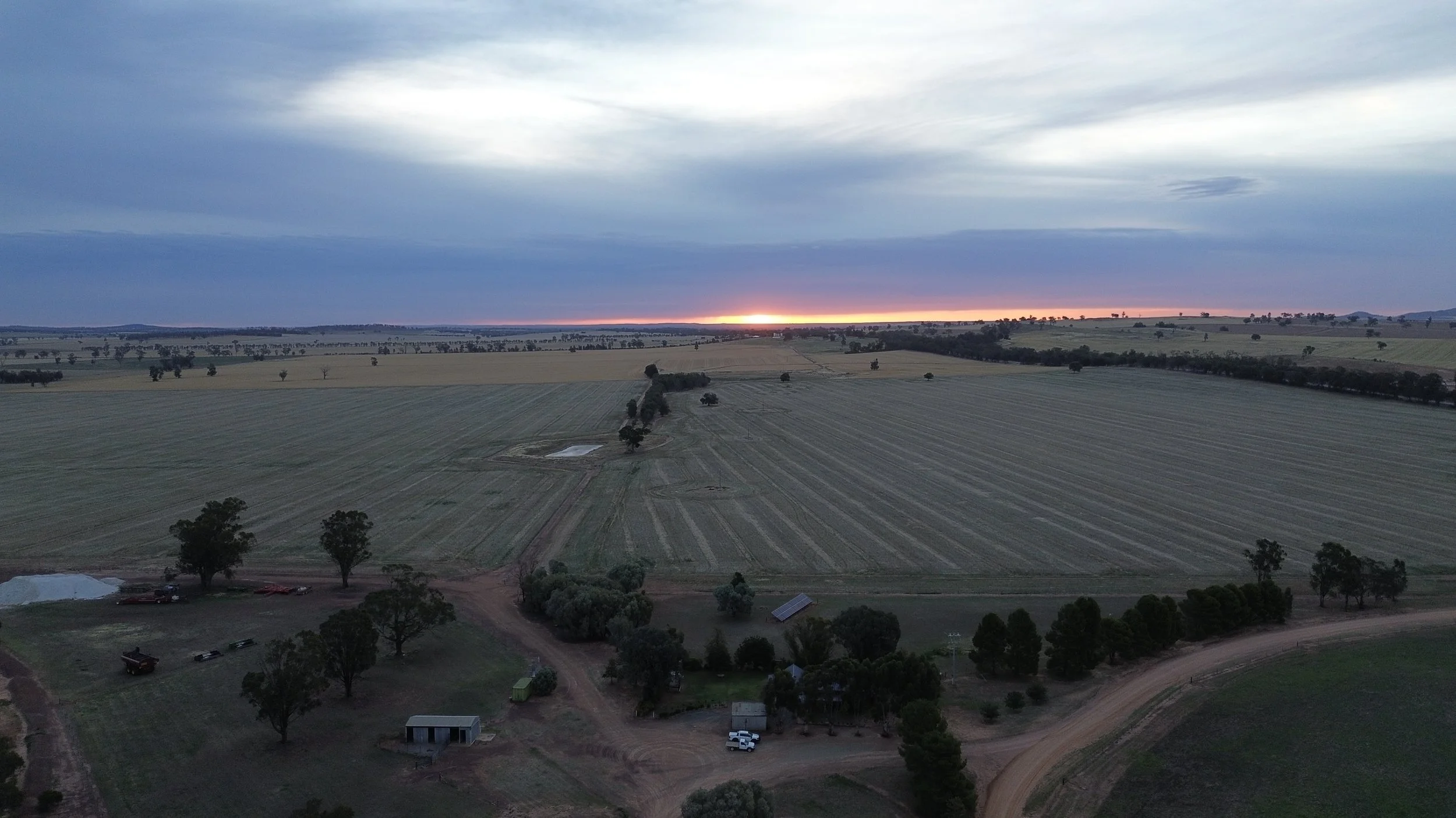 A vast rural farmland landscape at sunset with a cloudy sky, scattered trees, and dirt roads.