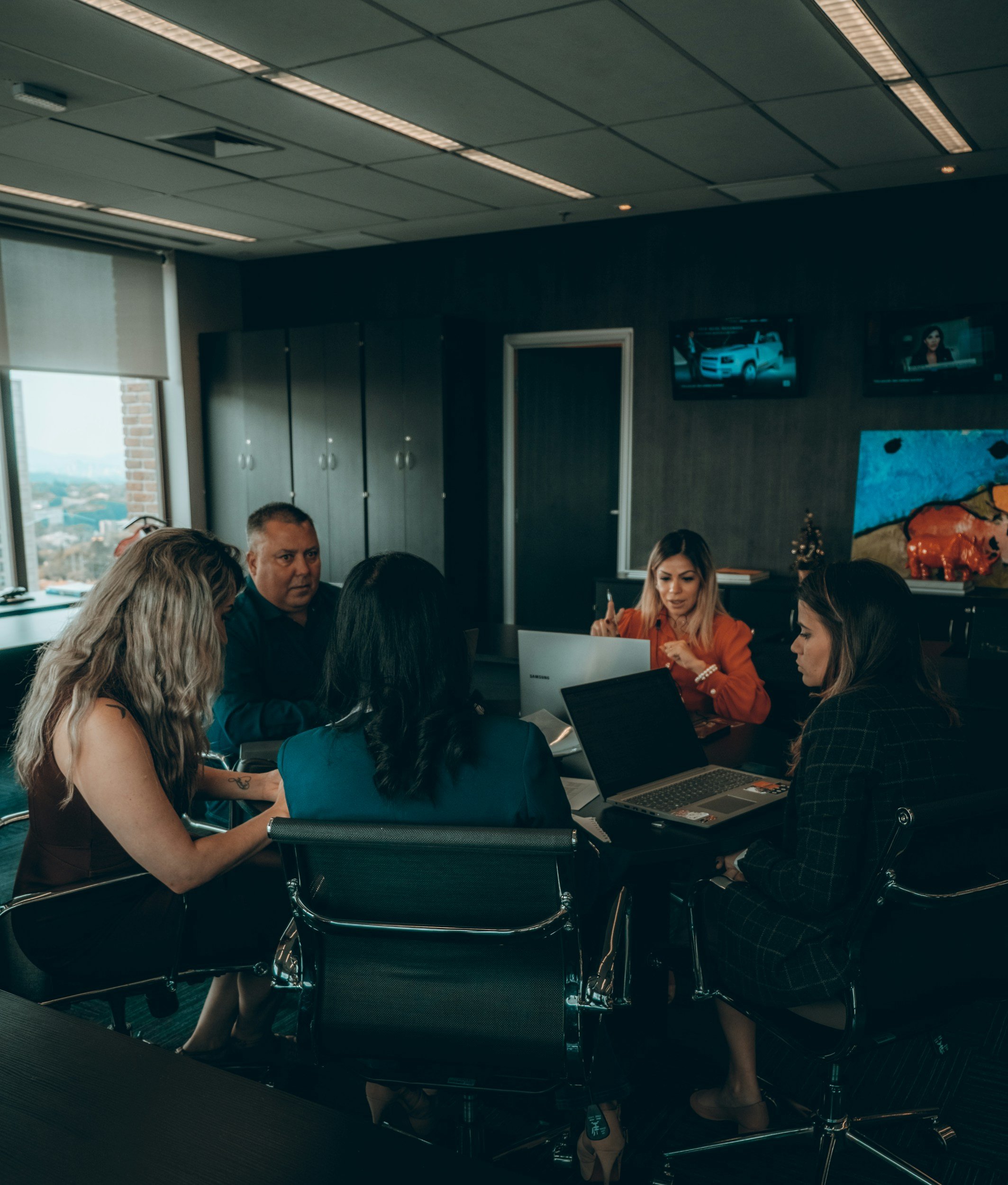 Business meeting in a modern office with five people seated around a table with laptops, discussing and collaborating.