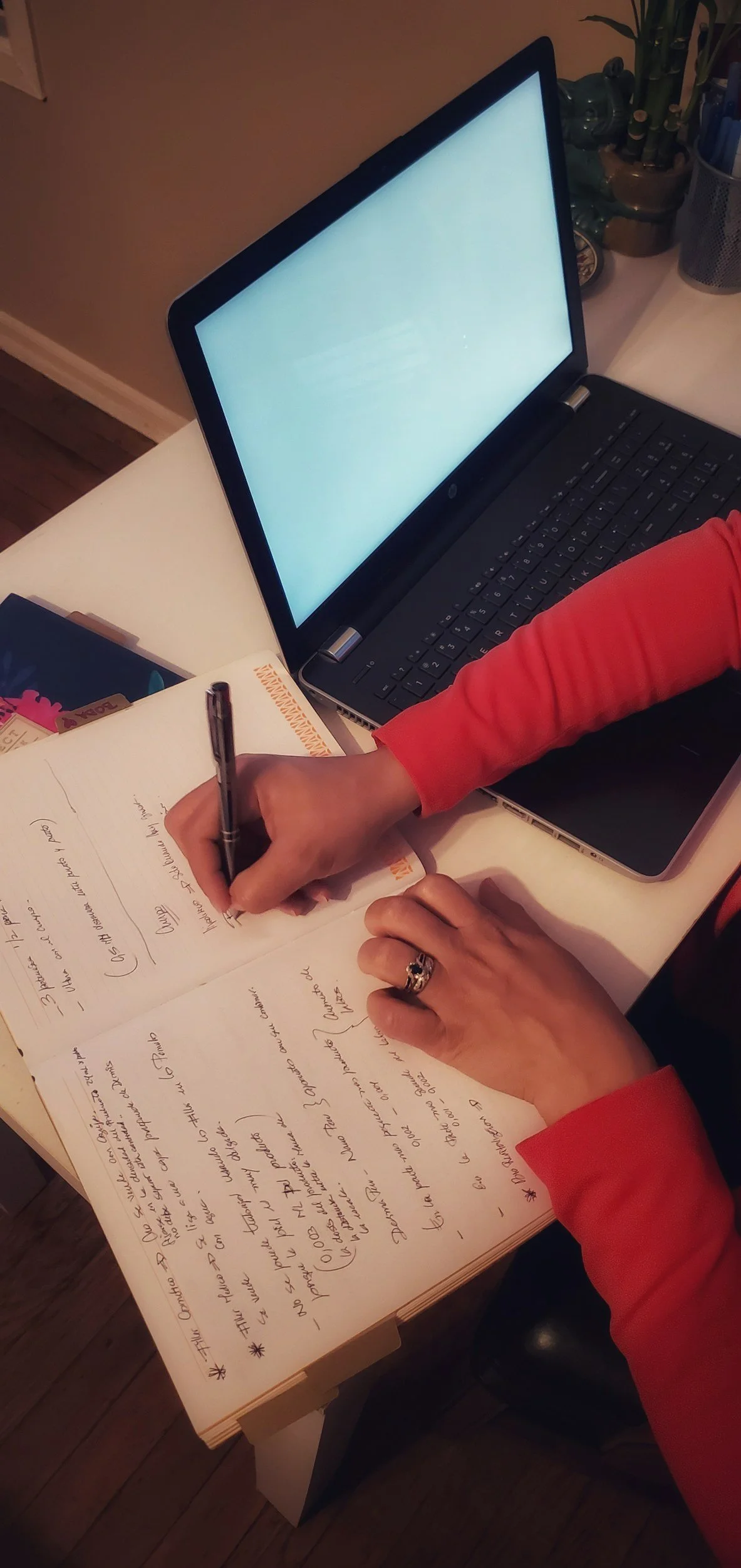 A person with a red long-sleeve shirt is writing in a notebook with handwritten notes, near a laptop with a blank screen, on a white table with some pens and a container of plants in the background.