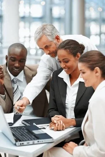 Business team in a meeting around a laptop, smiling and listening as a man points at the screen.