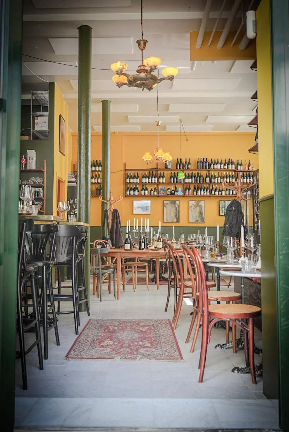 Indendørsareal af en restaurant med borddækning, vinflasker på hylder, og en gul væg med billeder.