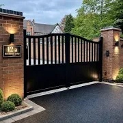 Black and brown retractable driveway gate between brick pillars with light fixtures, leading to a house, surrounded by trees and grass.