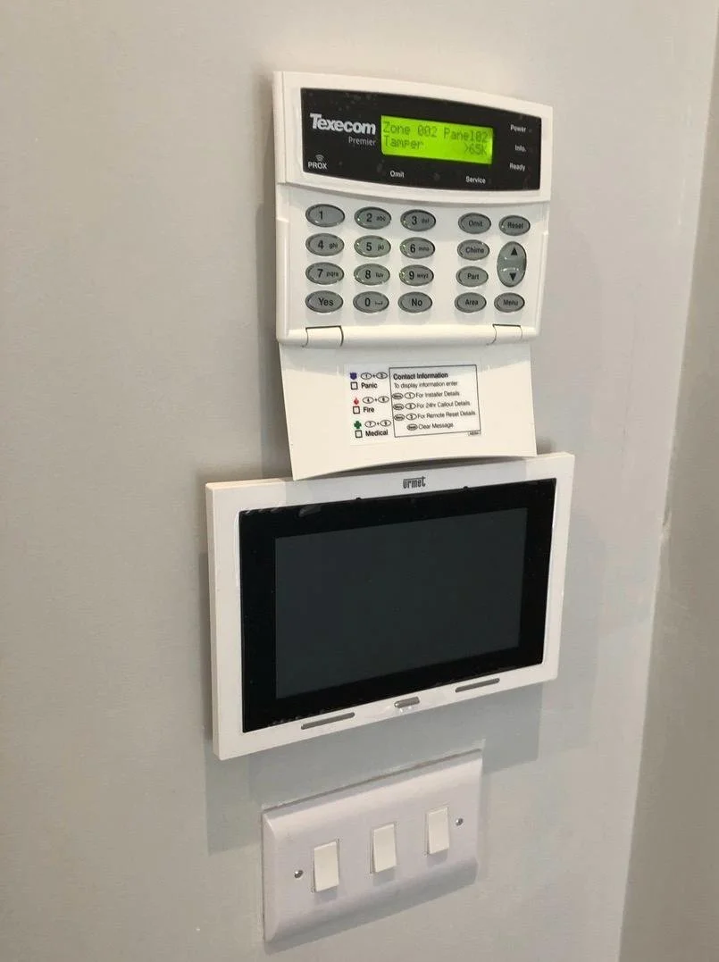 A security alarm control panel with a small screen displaying "Zone 002 Panel 02 Tamper." Below it is a white monitor, and below that are three electrical switches.