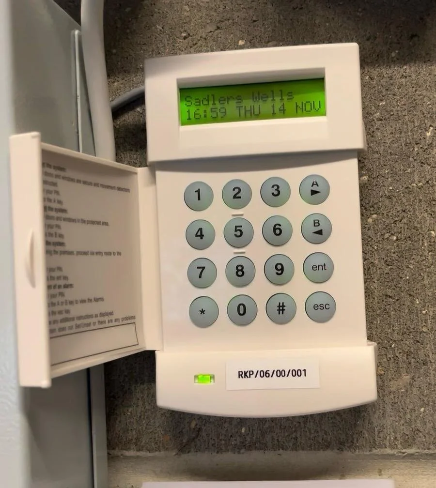 A white security keypad with an open door revealing instructions inside. The display reads 'Sadler's Wells 16:59 THU 14 NOV' and the keypad has numbered keys, arrows, 'ent,' 'esc,' '*' and '#' buttons. There is a small green light and a sticker with the code 'RKP/06/00/001'.