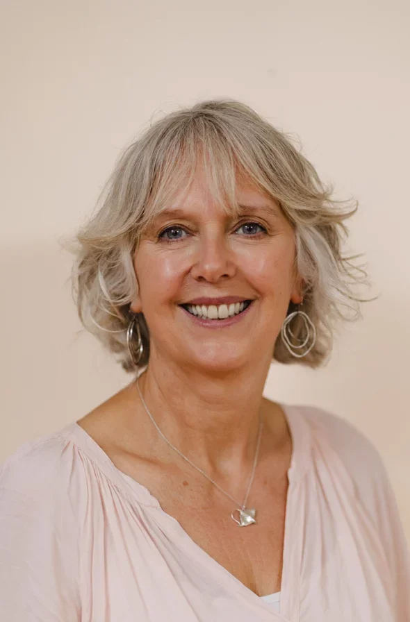 A middle-aged woman with short, wavy blonde hair smiling at the camera, wearing silver hoop earrings and a delicate silver necklace with a small pendant, dressed in a light pink blouse.