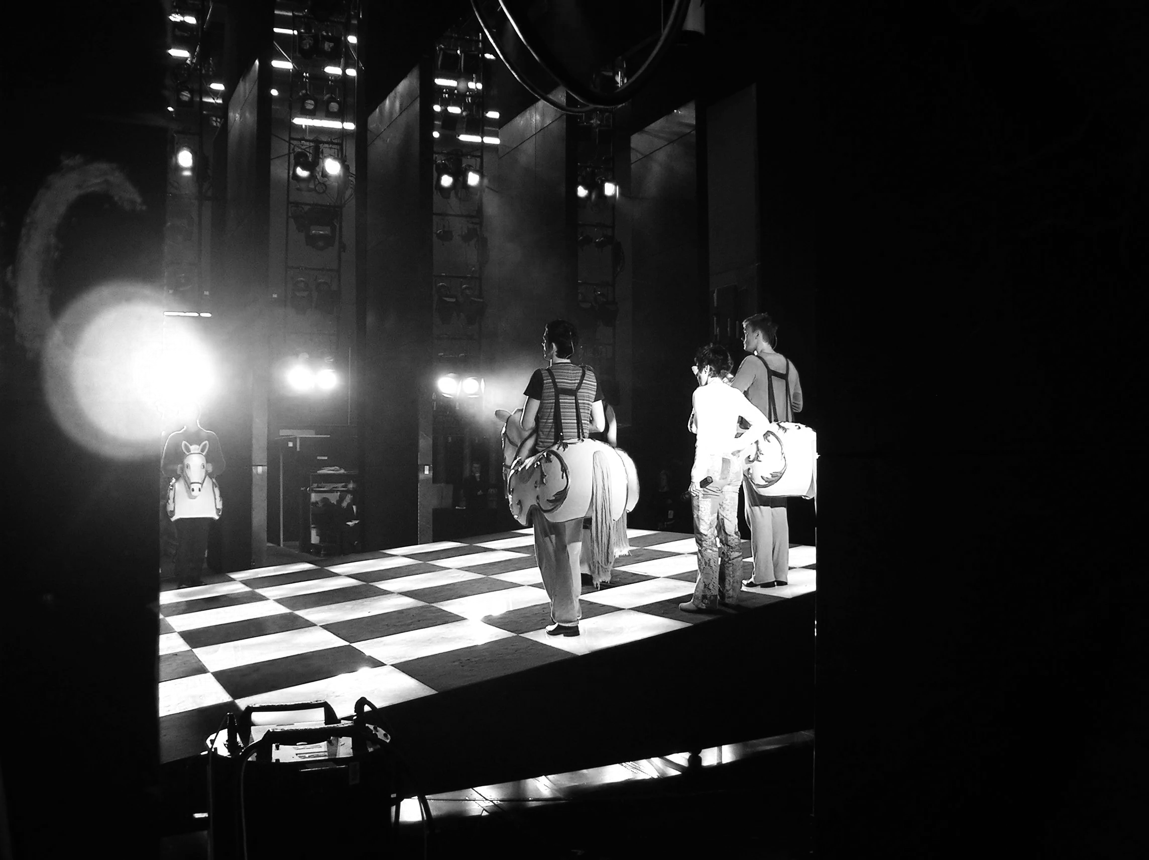 Schwarz-Weiß-Foto von einer Gruppe von Menschen, die vor einer Bühnenkulisse mit Scheinwerfern stehen, während sie auf eine Show oder Performance warten. Probenfoto