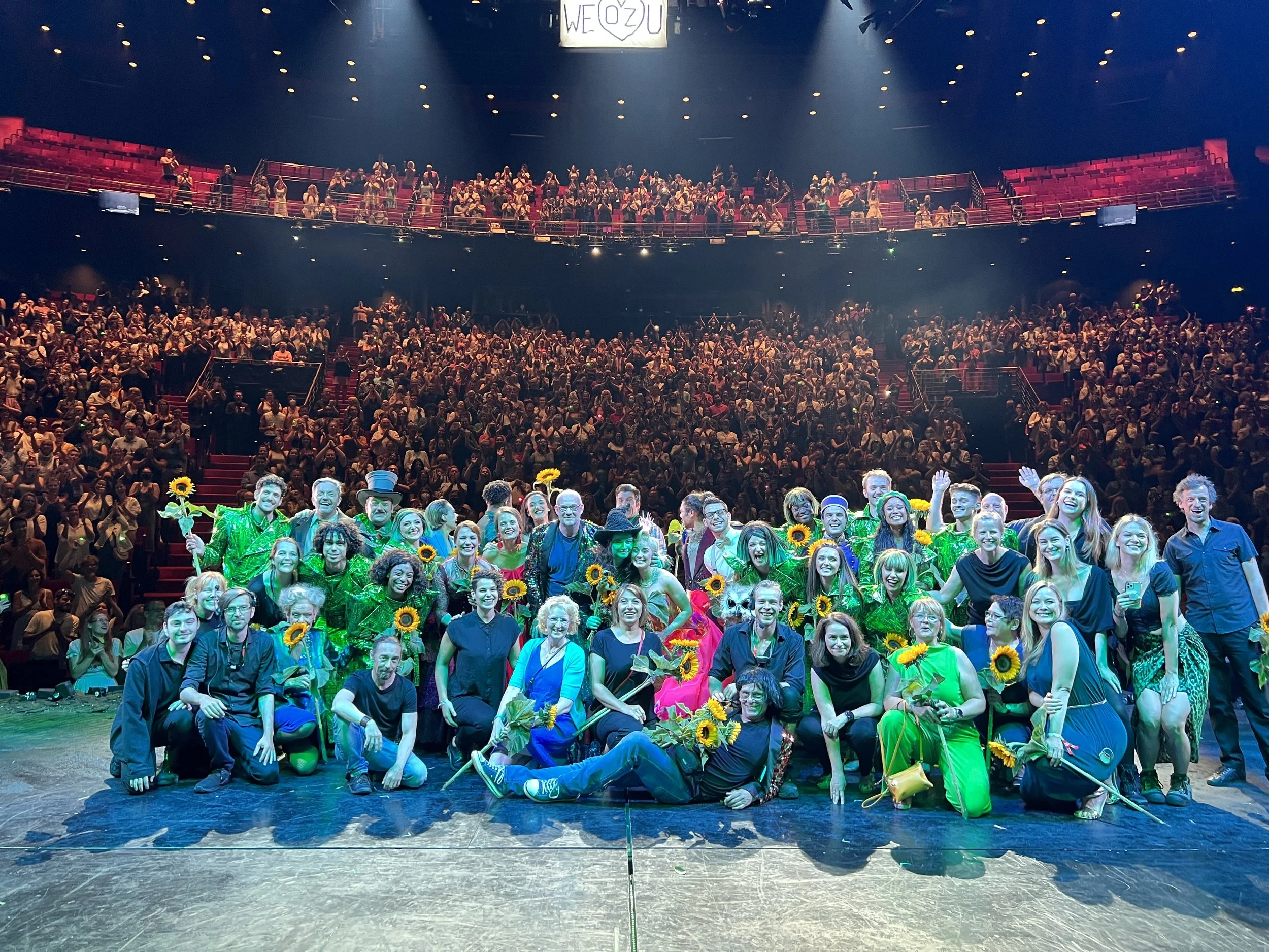Eine große Menschenmenge auf einer Bühne posiert für ein Gruppenfoto. Publikum sitzt im Theater. Im Vordergrund eine Gruppe lachender Menschen mit Sonnenblumen und bunten Kostümen. Abschiedsvorstellung von Wicked - die Hexen von Oz