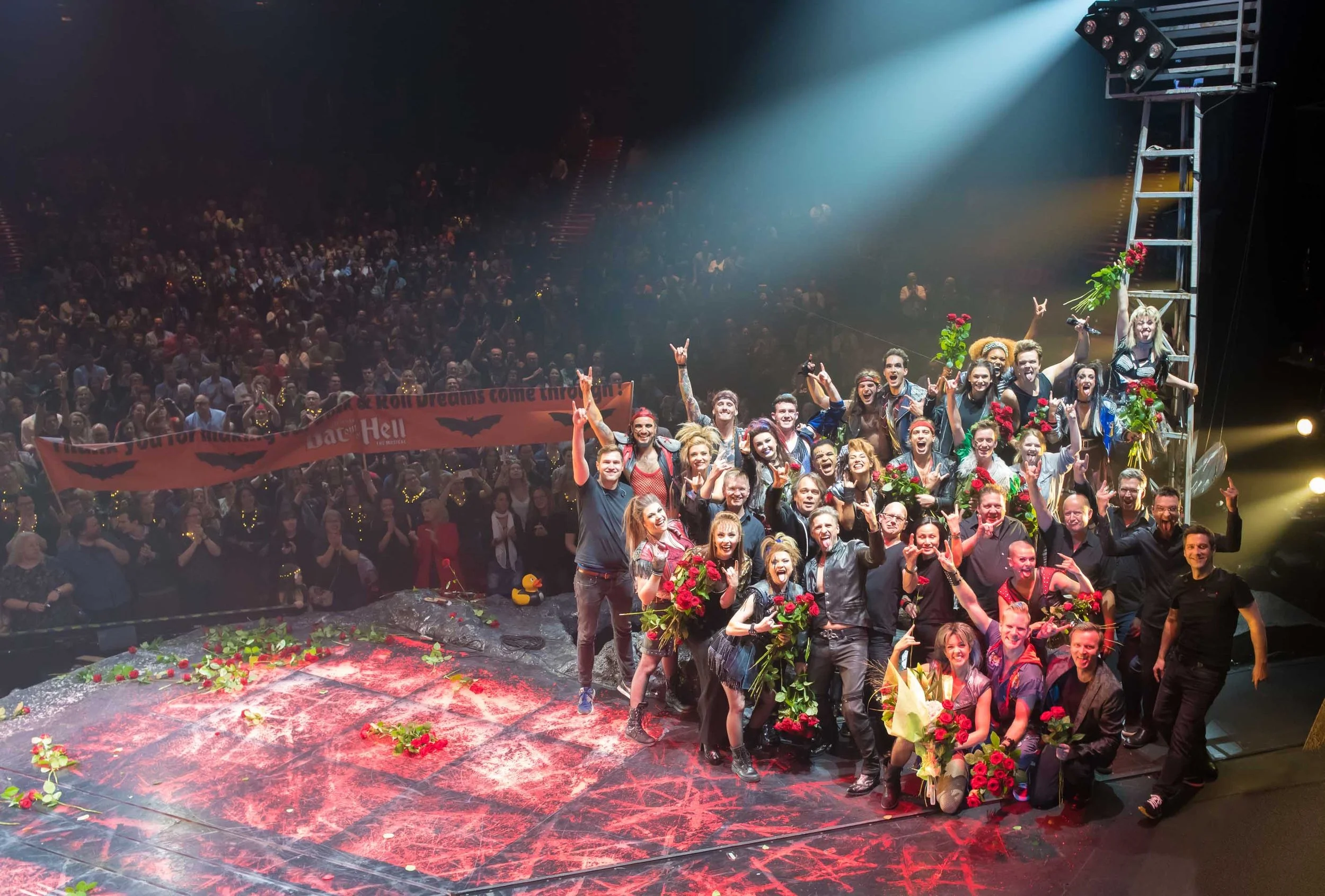 Gruppe von Menschen auf einer Bühne, die Blumen und Jubel zeigen, während das Publikum im Hintergrund zuschaut, bei einer Festveranstaltung mit Halloween-Dekorationen auf dem Boden.