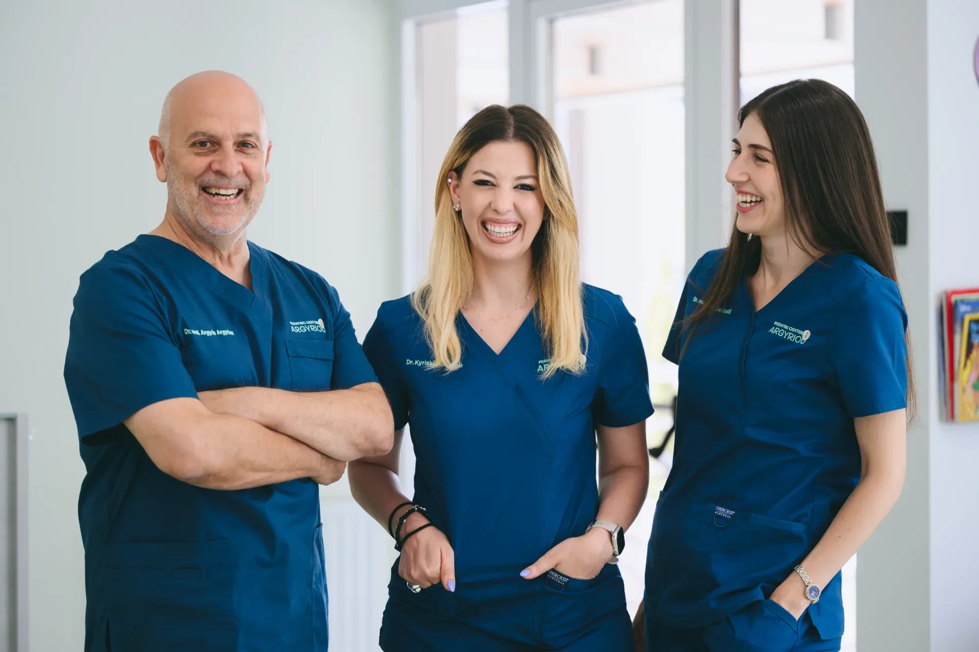 Three healthcare professionals wearing blue scrubs, smiling and standing together indoors near a window.