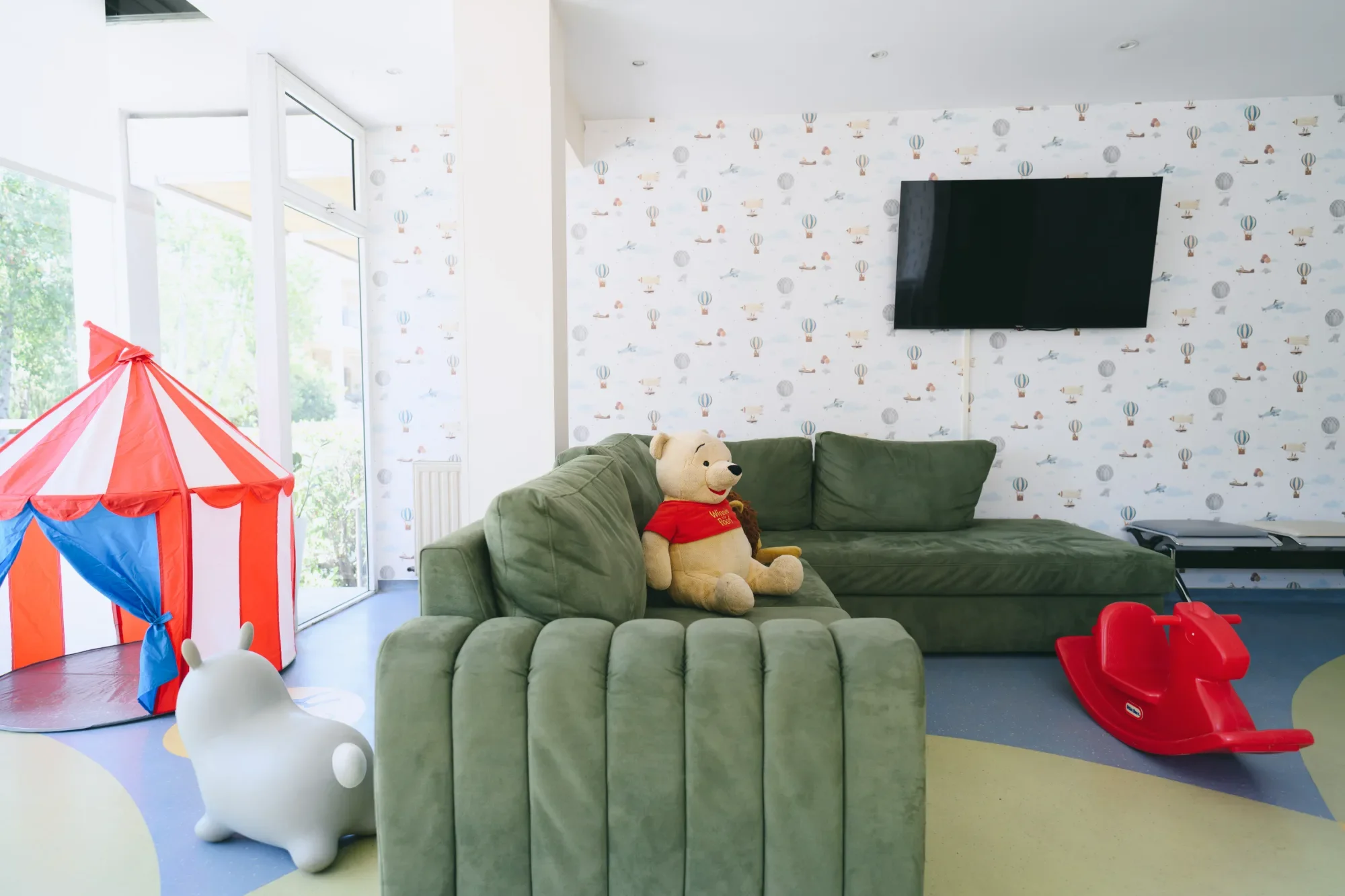 Children's playroom with a red and white striped circus tent, a white bunny-shaped ride-on toy, a green sofa with Winnie the Pooh plush toy, a mounted flat-screen TV, colorful wallpaper with airplane and hot air balloon patterns, and a red plastic sled.