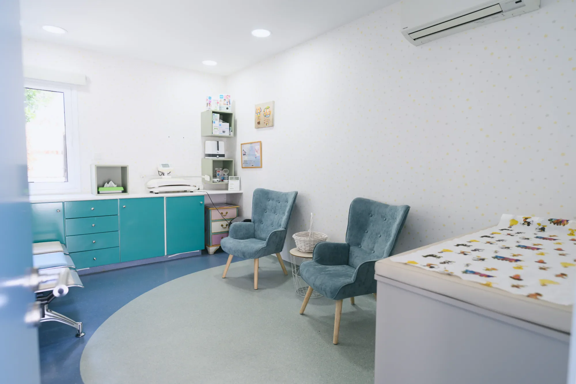 Hospital or clinic room with blue and gray furniture, chairs, and medical equipment, featuring a window and decorative wall art.