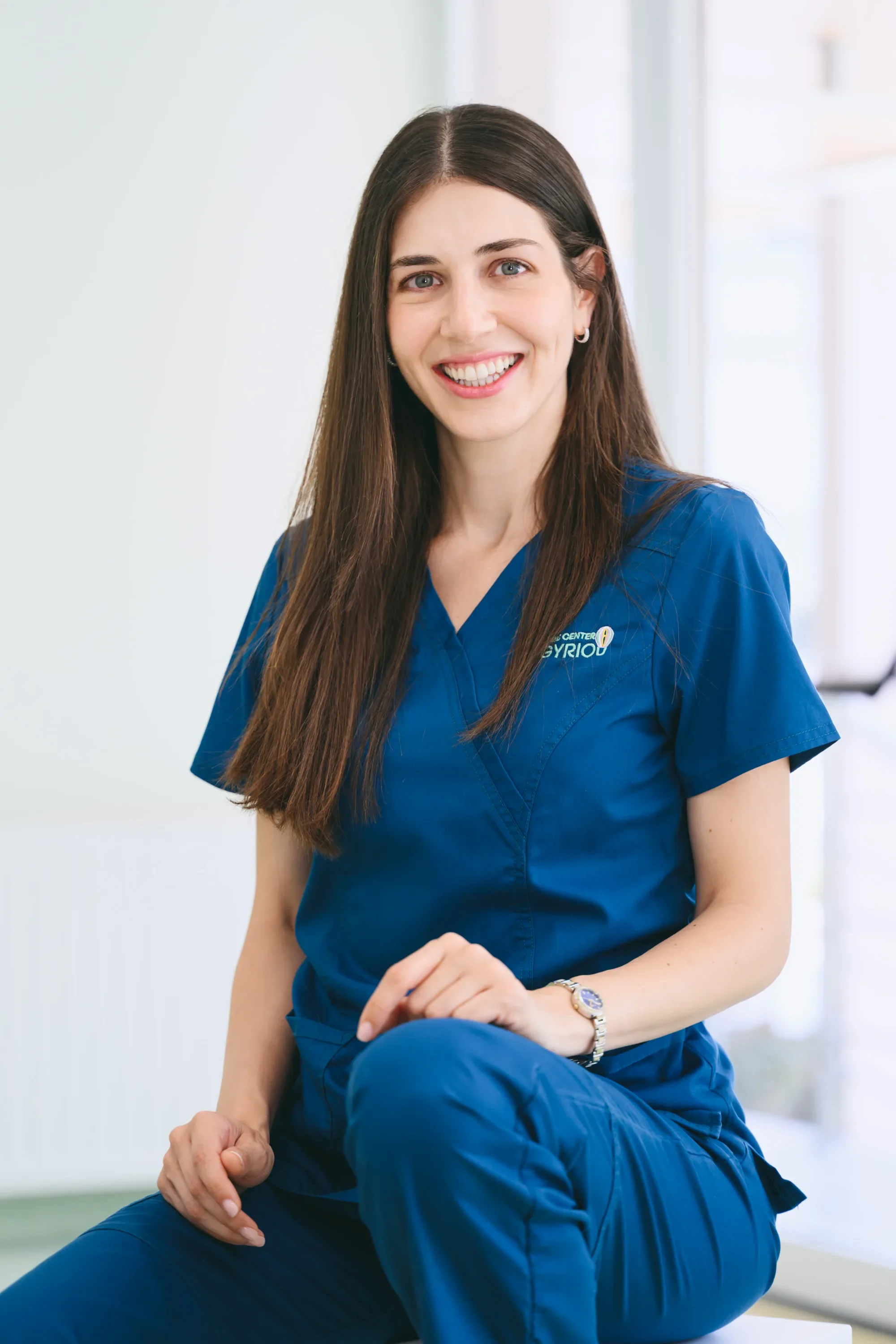 A young woman doctor with long dark hair, wearing blue medical scrubs, sitting in a medical setting, smiling at the camera.