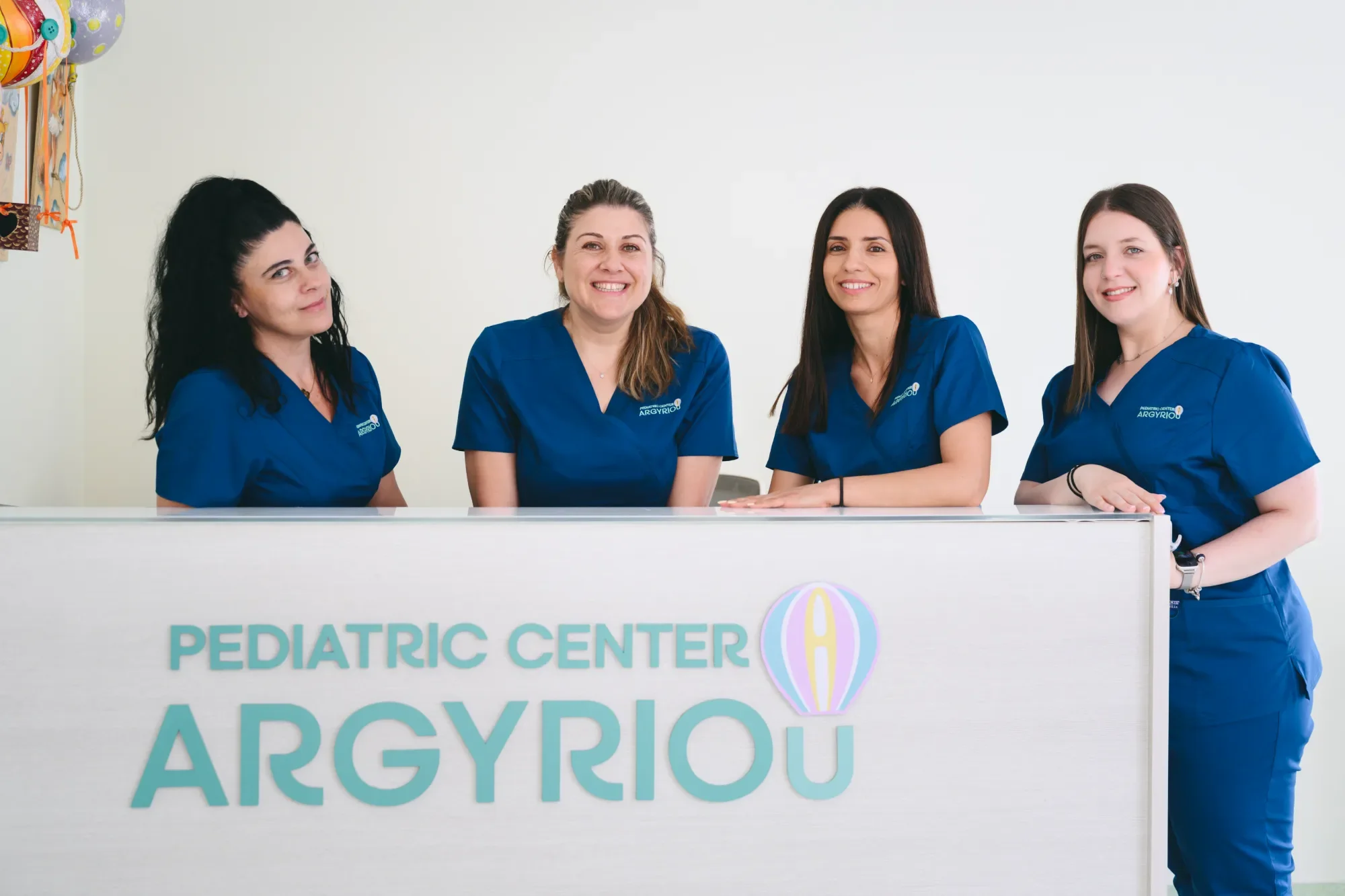 Four women in blue scrubs sitting and standing at a reception desk for a pediatric center, smiling at the camera.