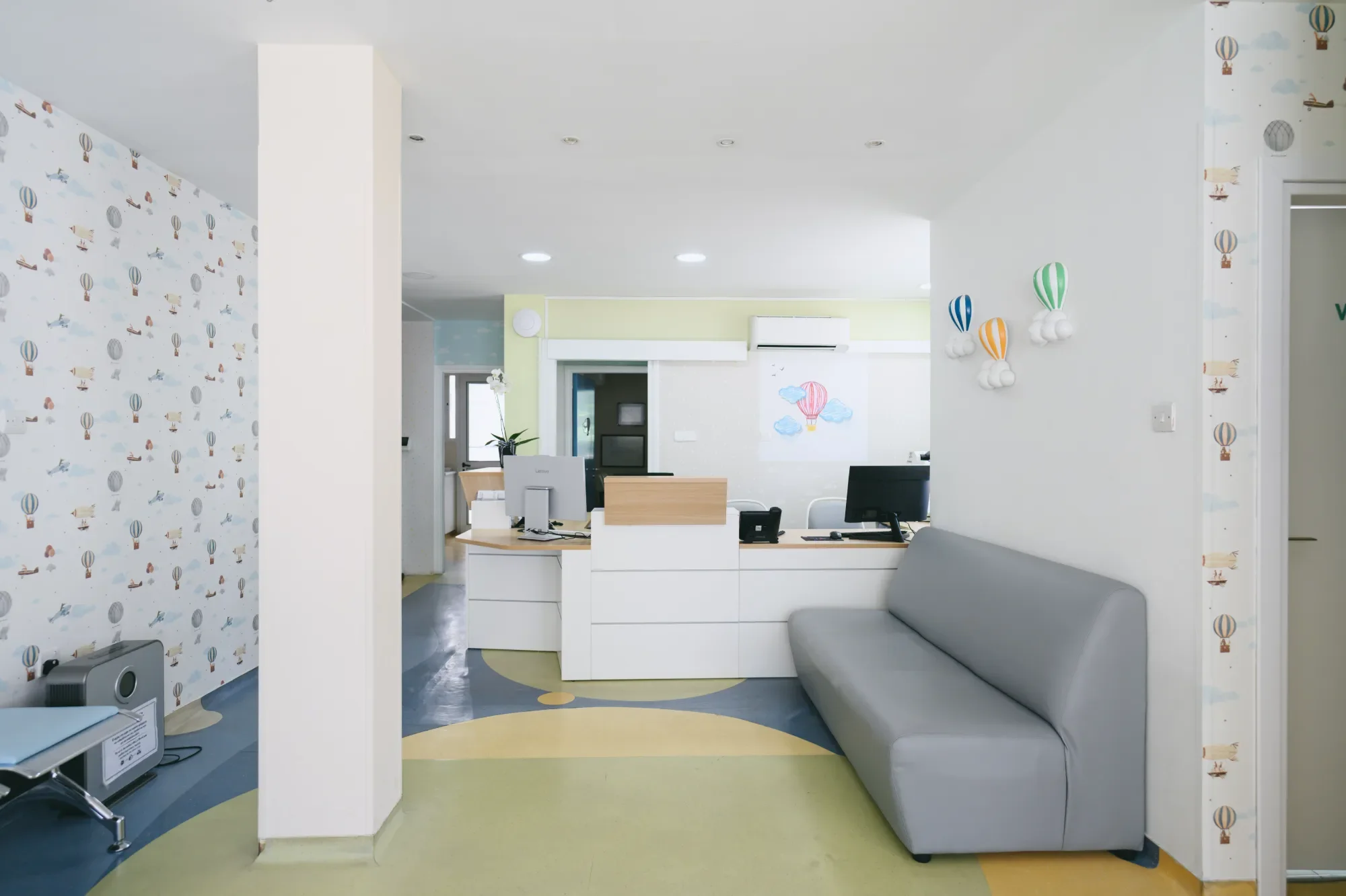 A reception area in a children's clinic decorated with hot air balloon-themed wallpaper and 3D hot air balloon wall art. There is a white reception desk, a gray sofa, and a colorful circular rug.