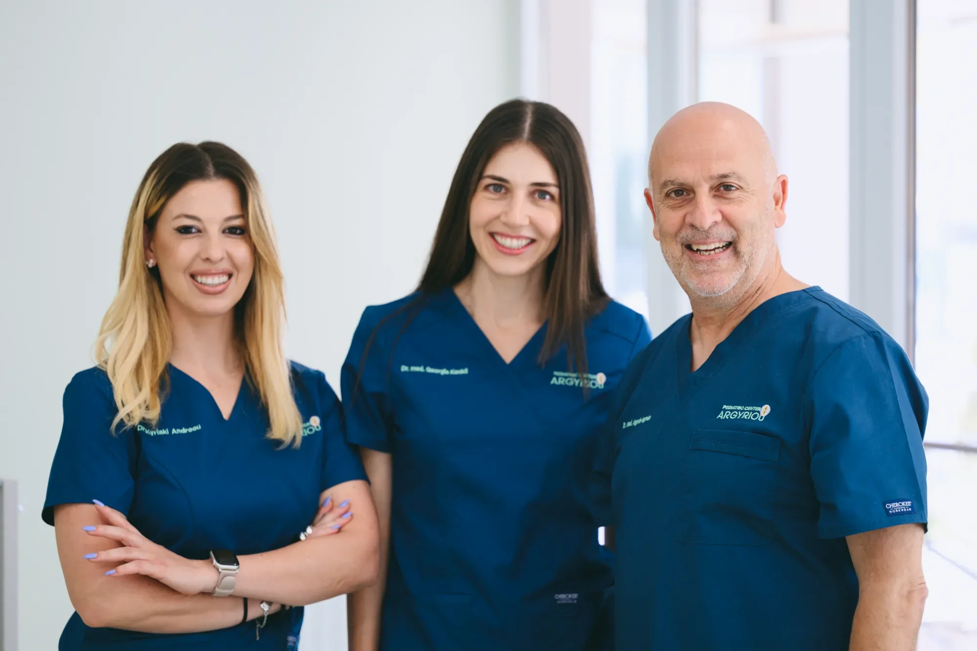 Three medical professionals in blue scrubs smiling and standing together in a hospital or clinic setting.