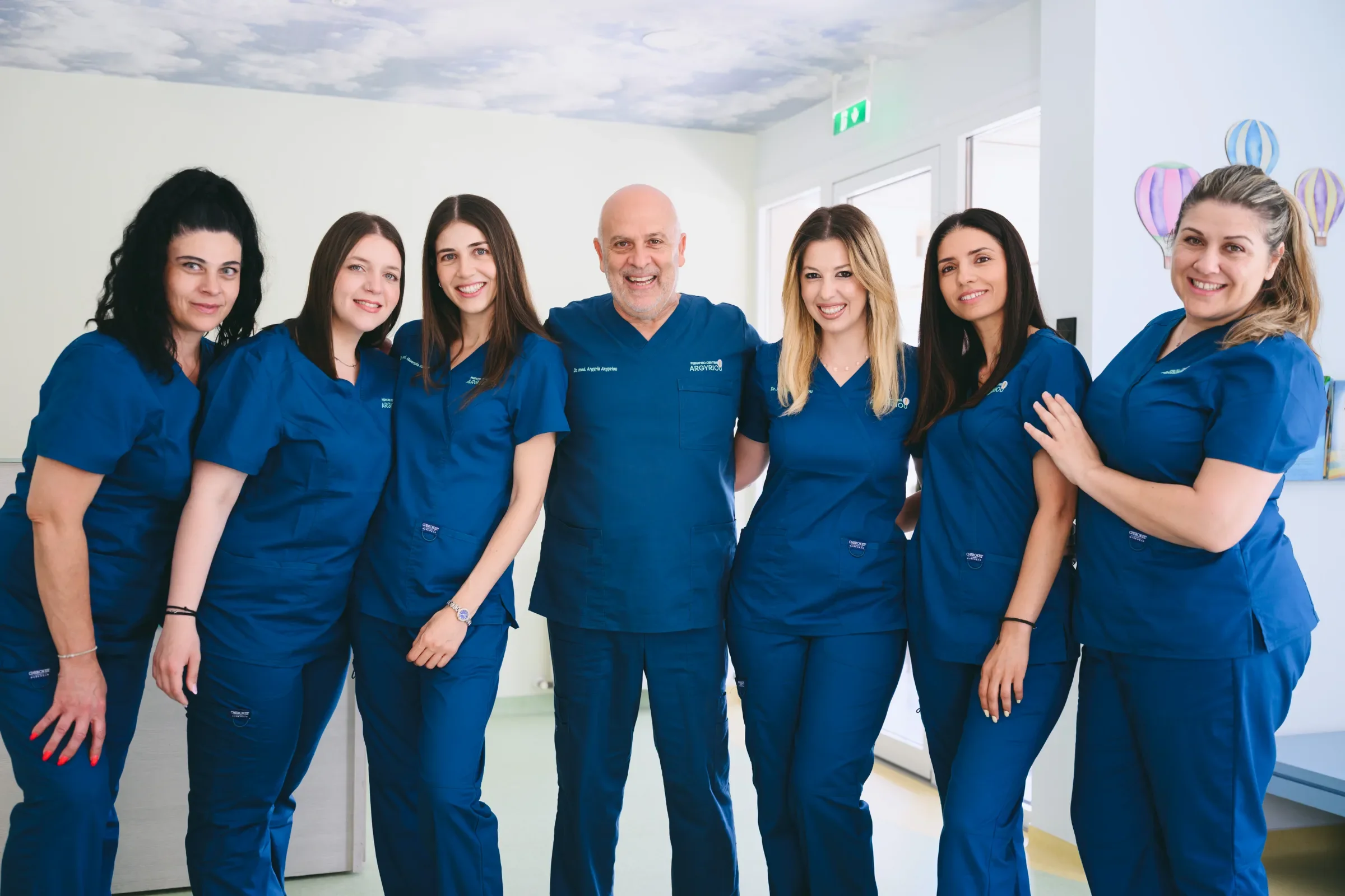 Group of seven healthcare professionals wearing blue scrubs, standing together indoors, smiling at the camera.