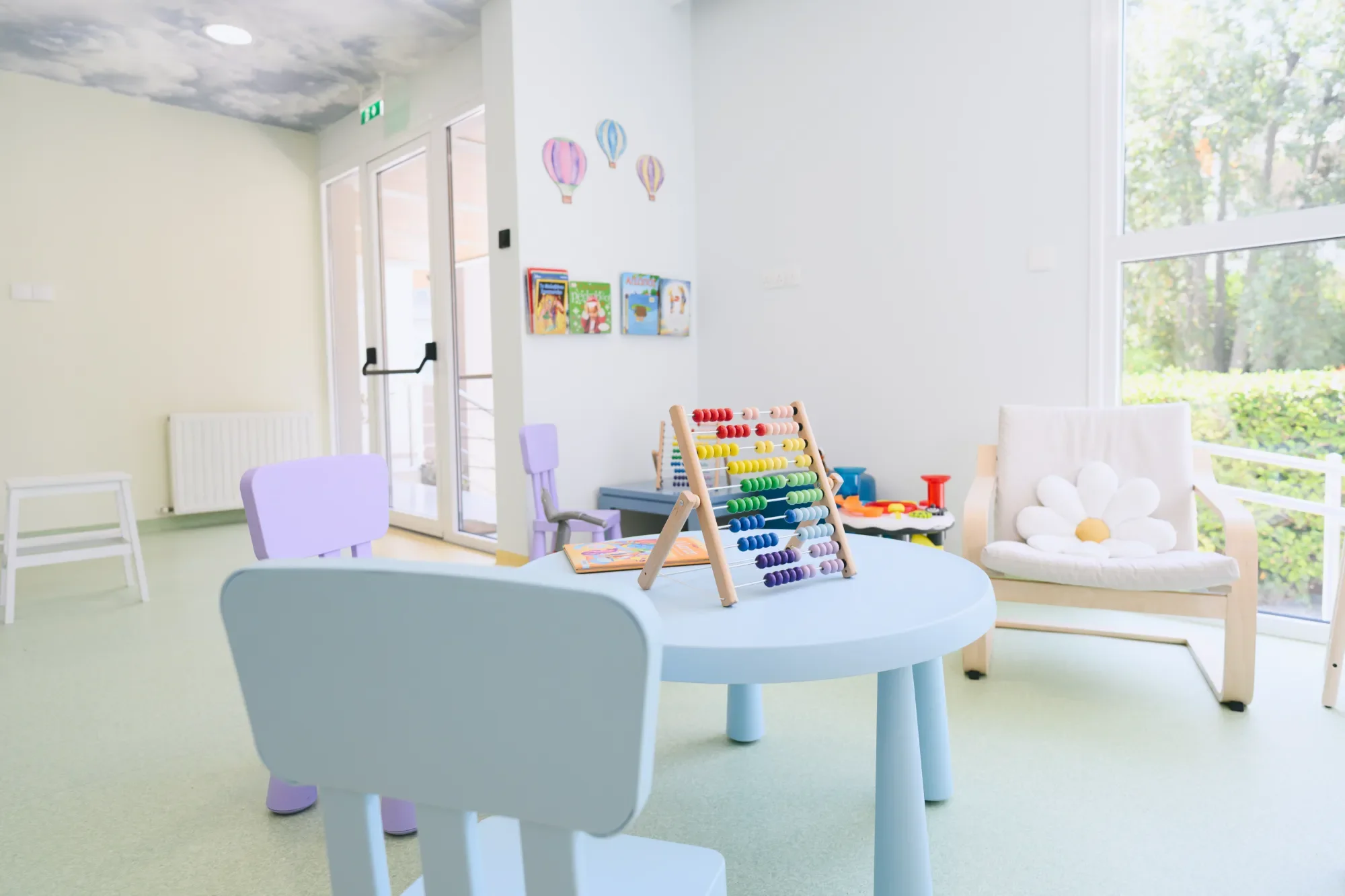 Colorful toys and books on a table in a bright children's playroom with pastel-colored chairs, large windows, and wall decorations of hot air balloons.