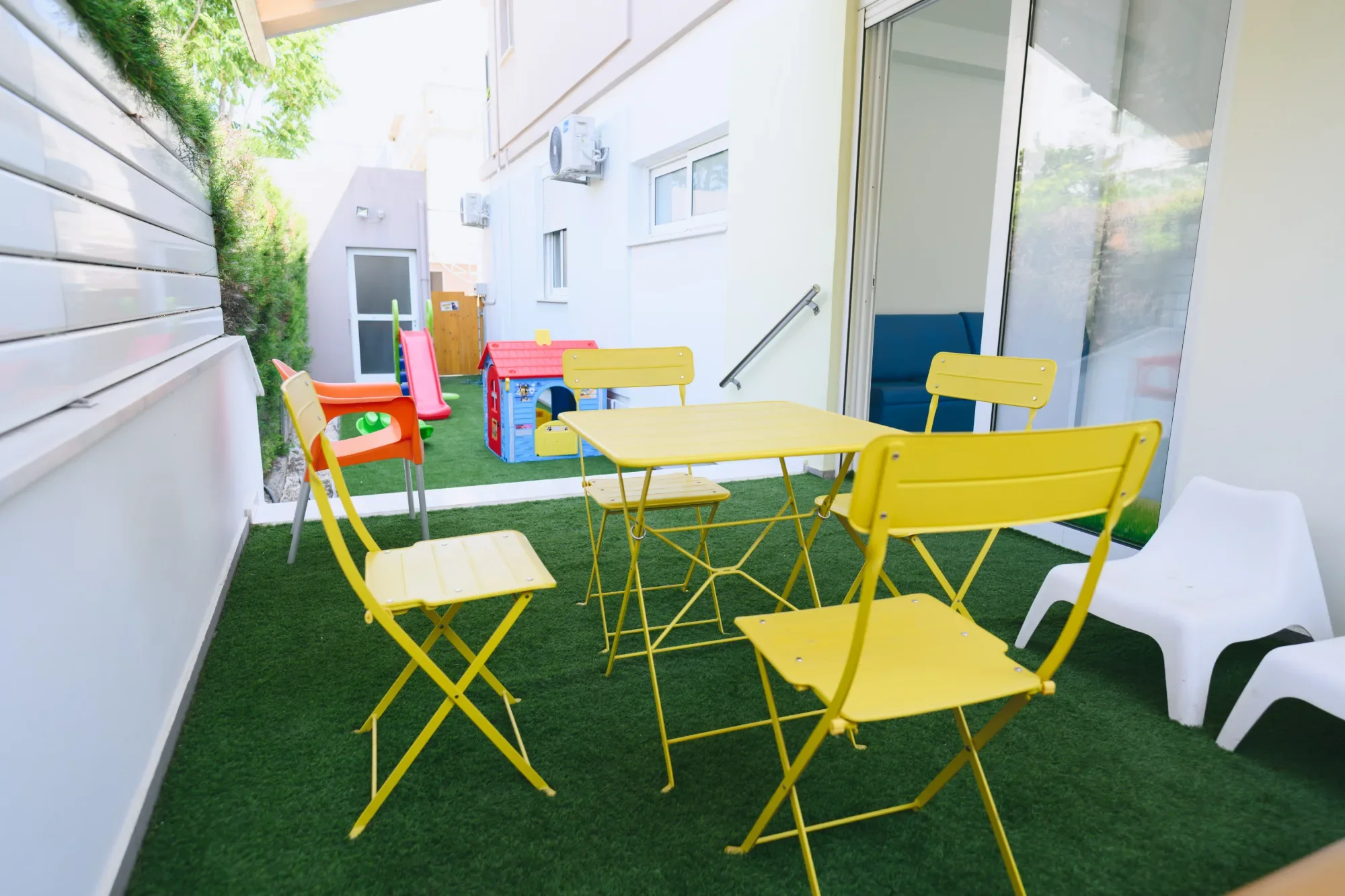 A small outdoor play area with yellow chairs and table, a playhouse, a slide, and other colorful toys, enclosed by white walls with a grassy floor.