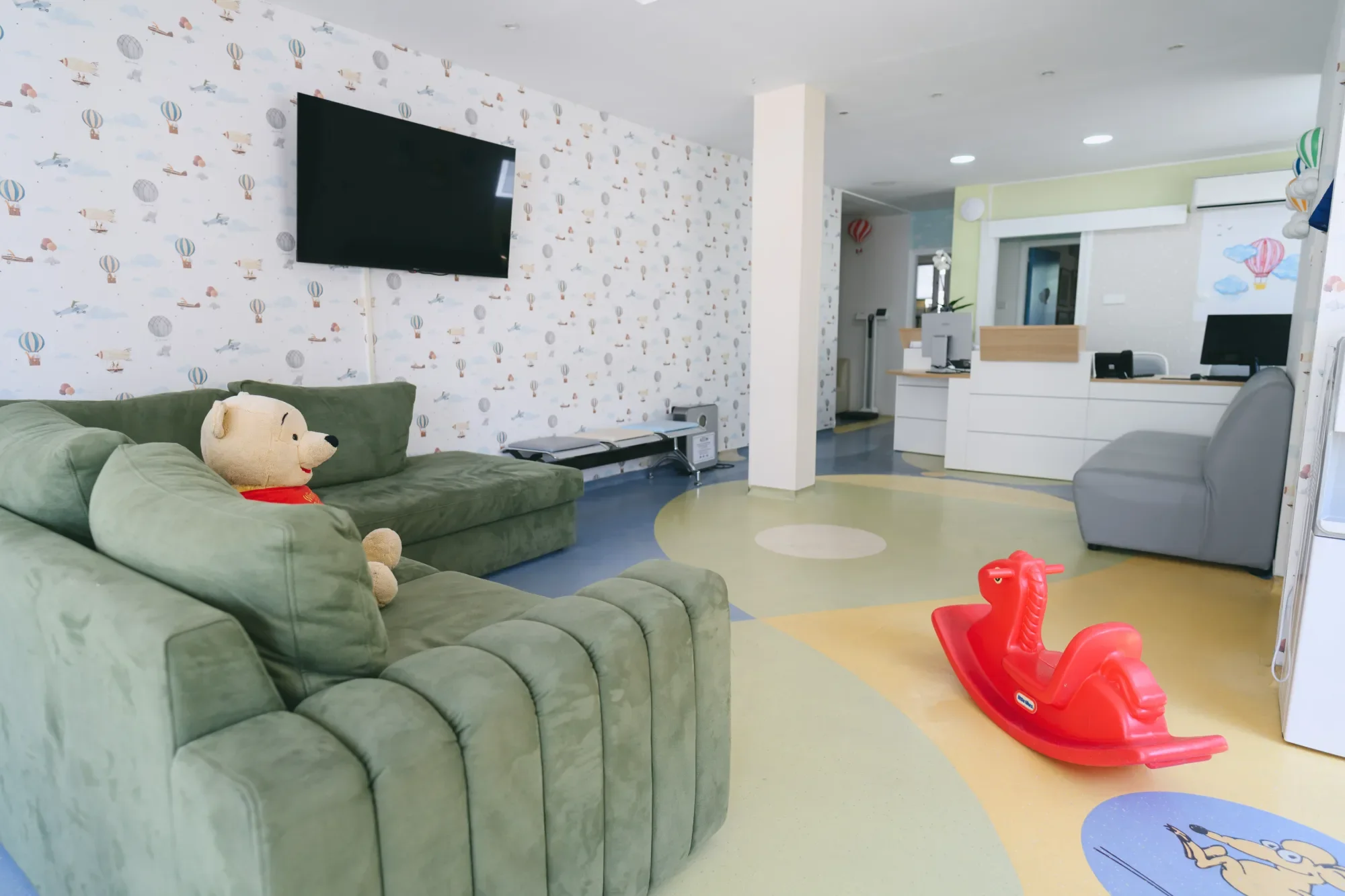 Child's play area with green sofa, a teddy bear, a red rocking horse, and a colorful floor. An entertainment area with a black TV on the wall and a computer workstation in the background.