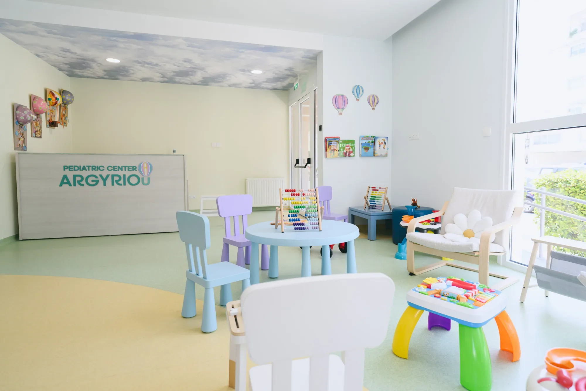 Bright, colorful pediatric center room with small chairs, toys, and books for children, large windows, and playful ceiling with cloud design.