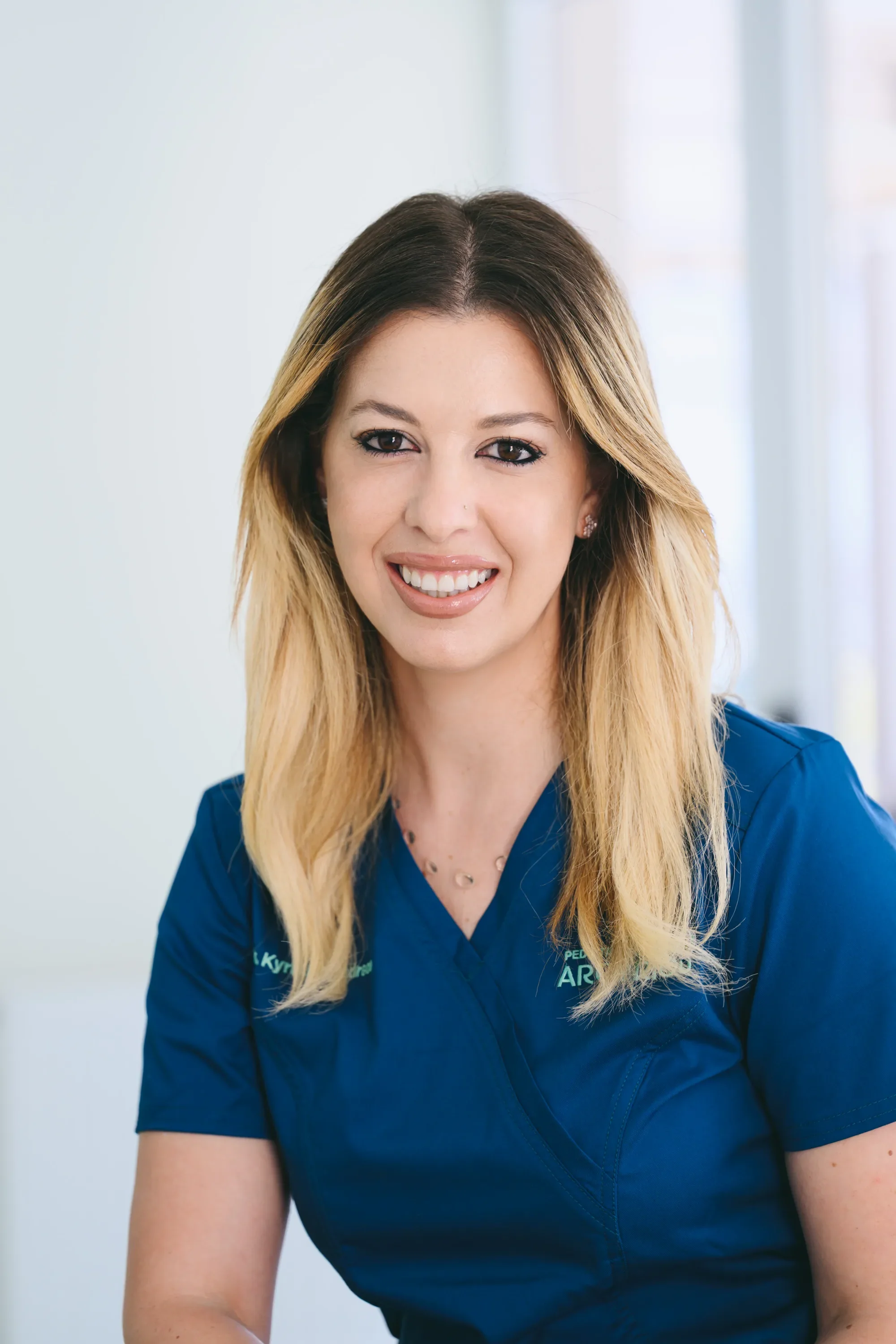 A woman doctor with long blonde hair smiling in a blue medical uniform.
